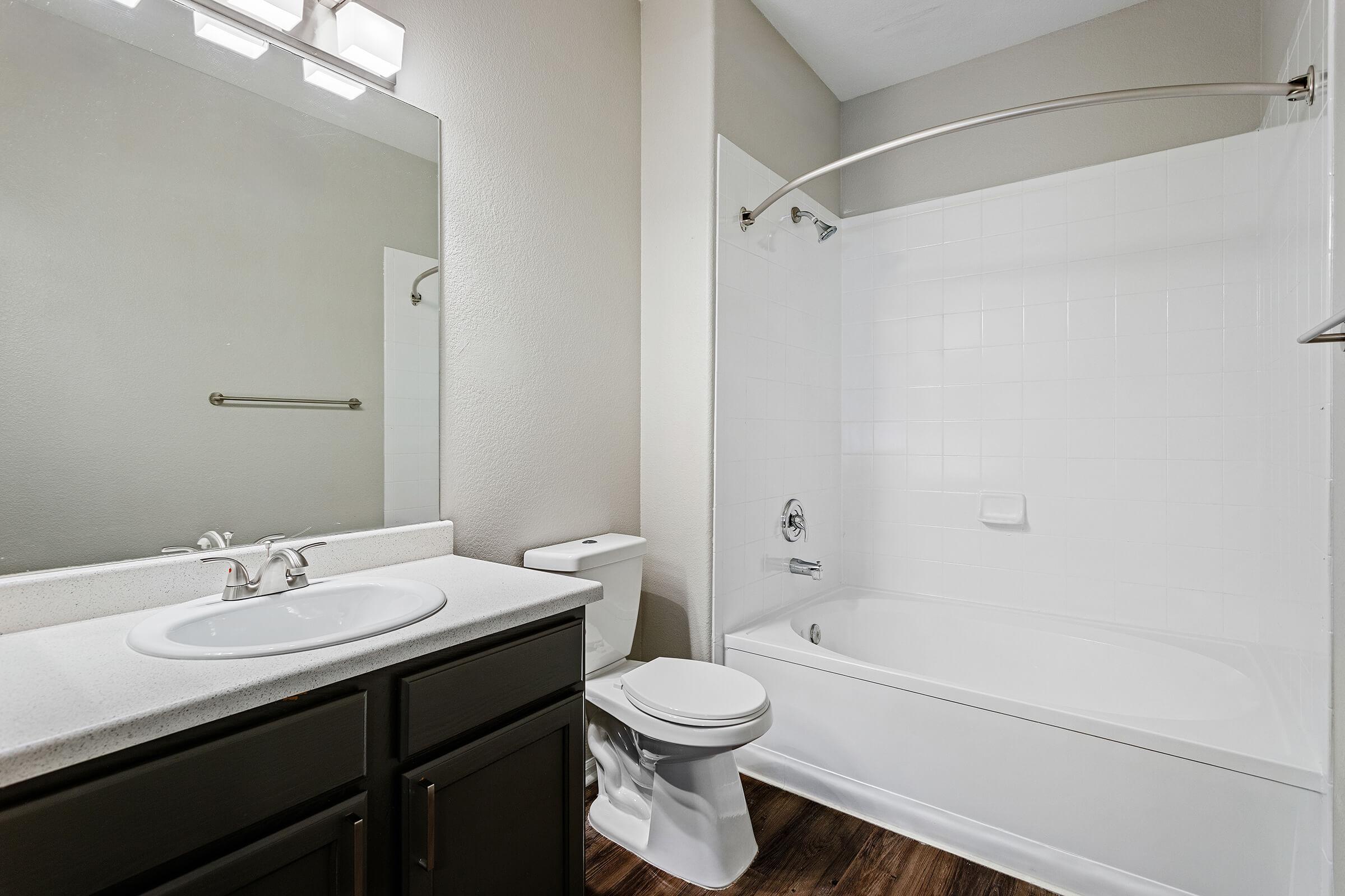 Bright and modern bathroom featuring a white tub and shower combo, a sleek mirror above a dark wood vanity with a sink, and a white toilet. The walls are a soft neutral color, and the flooring appears to be wood-like laminate. A chrome towel bar is mounted on the wall.