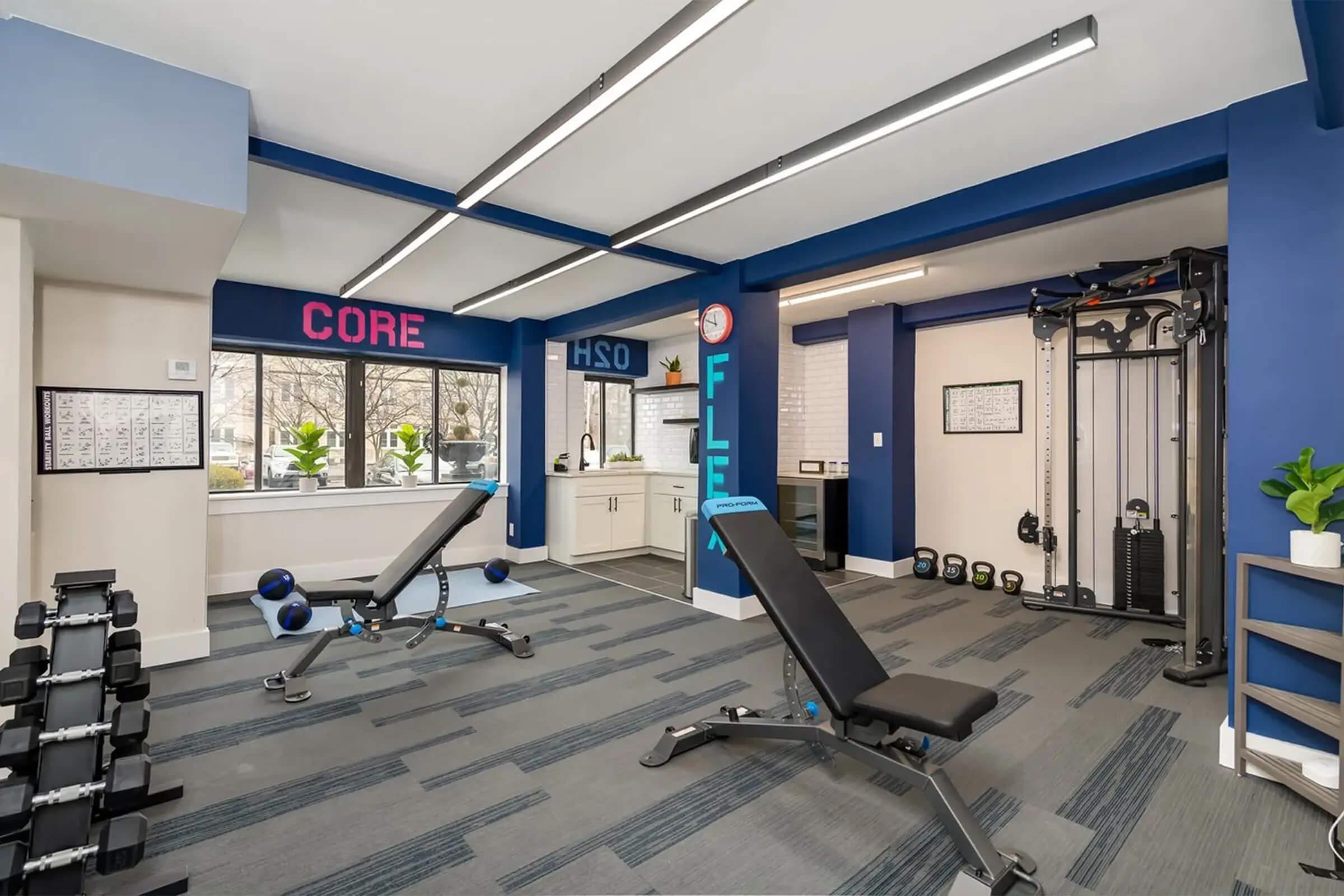 A modern fitness room featuring weight benches, free weights, and a cable machine. Bright blue walls adorned with motivational words like "CORE" and "FLEX." Large windows provide natural light, and potted plants add a touch of greenery to the space.