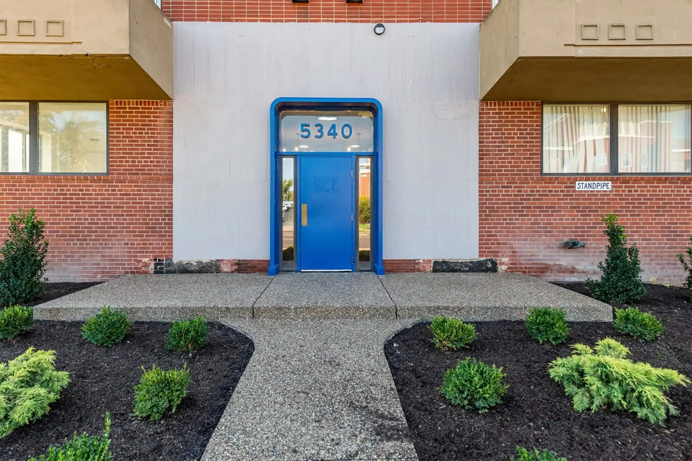 A modern apartment building entrance with a bright blue door displaying the number 5340. The pathway is lined with small shrubs and fresh landscaping. The facade features a combination of brick and smooth walls, with windows on either side of the door. The overall appearance is clean and inviting.