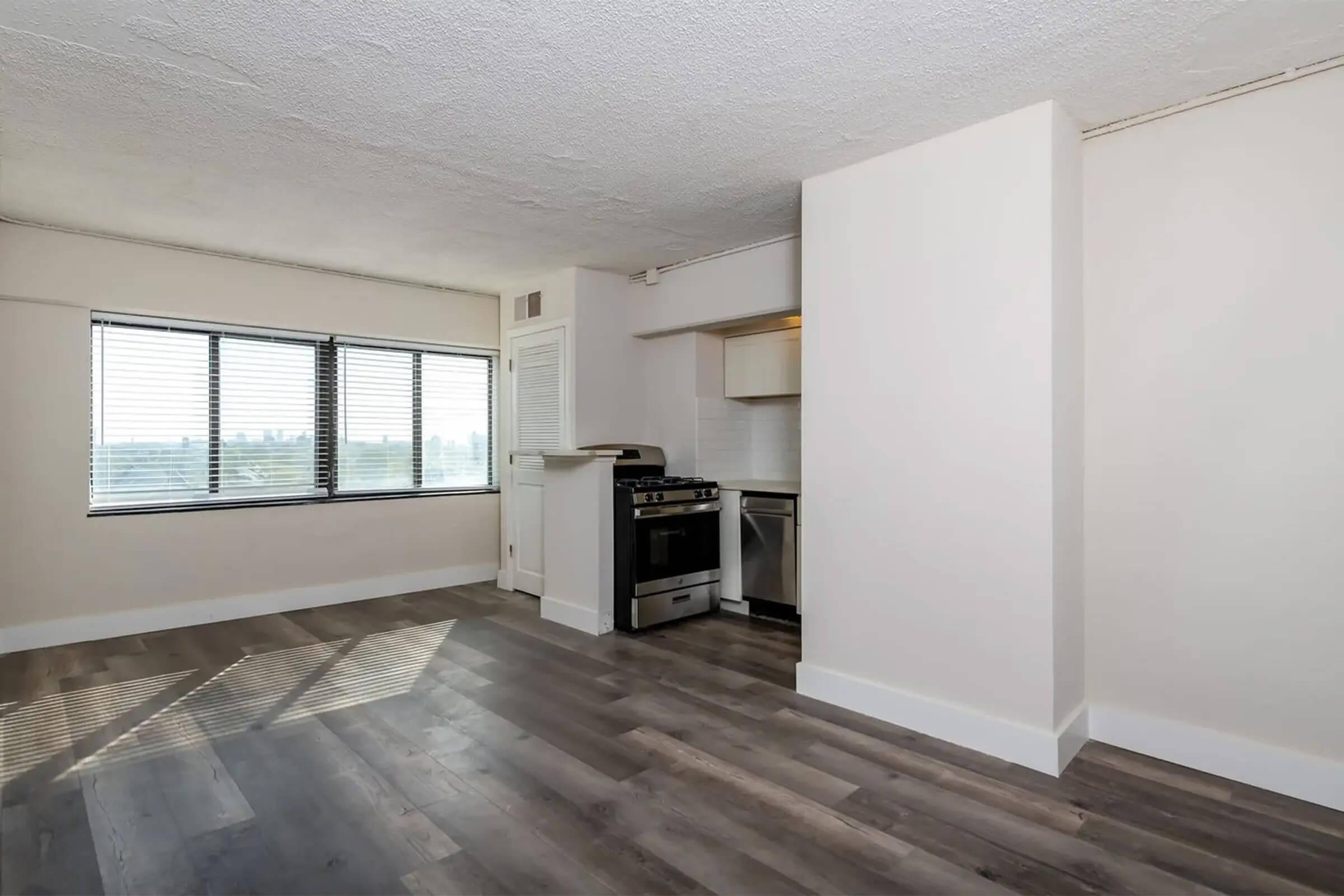A spacious, modern apartment interior featuring large windows with blinds, hardwood-style flooring, and a small kitchen area with a stainless steel stove and refrigerator. The walls are painted in a light color, creating an airy feel. Natural light fills the space, highlighting its open layout.