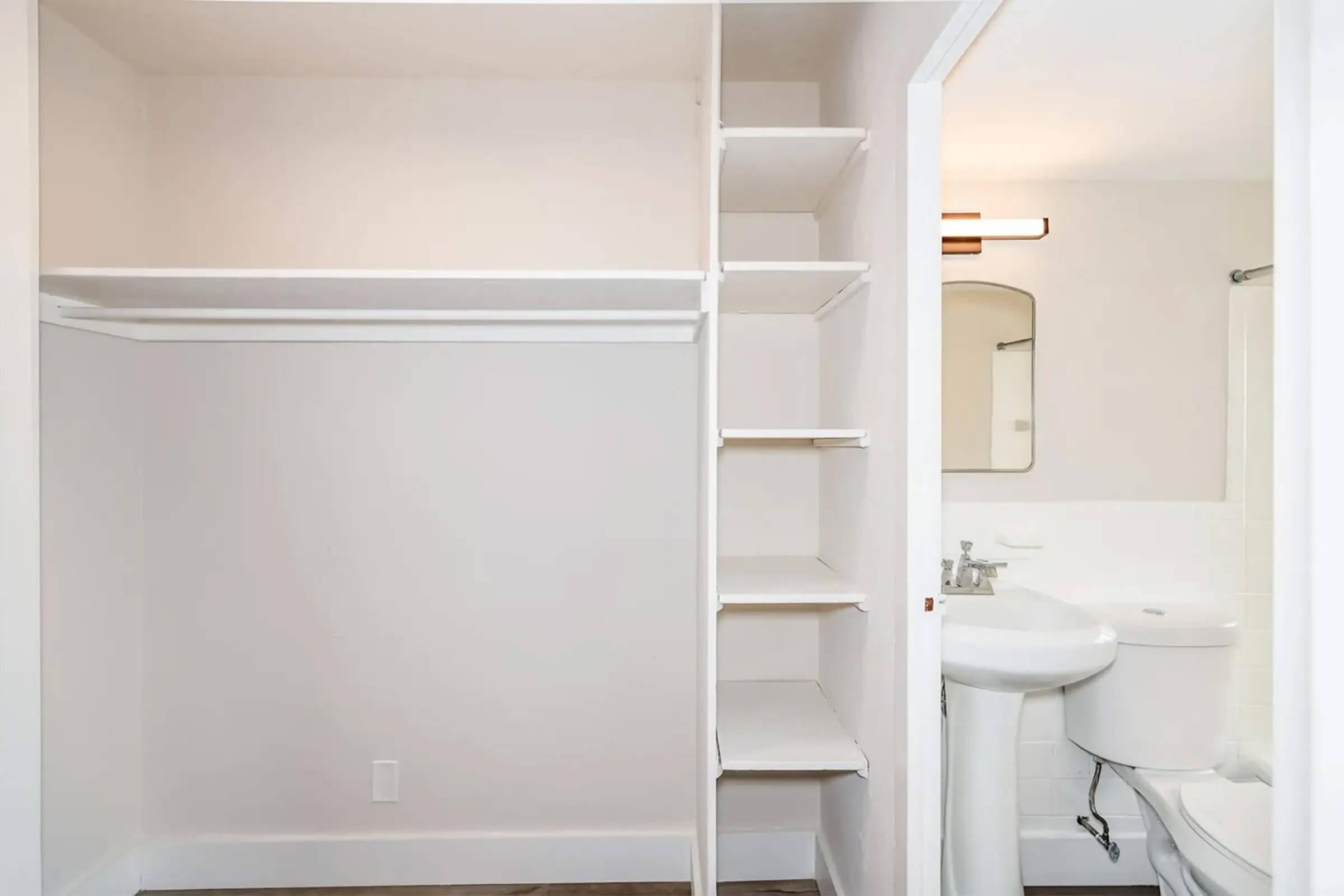 A view of a minimalist bathroom and closet area. The closet features open shelving and a hanging rod, while the bathroom includes a small sink and mirror. The color scheme is light and neutral, creating a clean and spacious feel. Natural light comes into the area, enhancing the overall brightness.