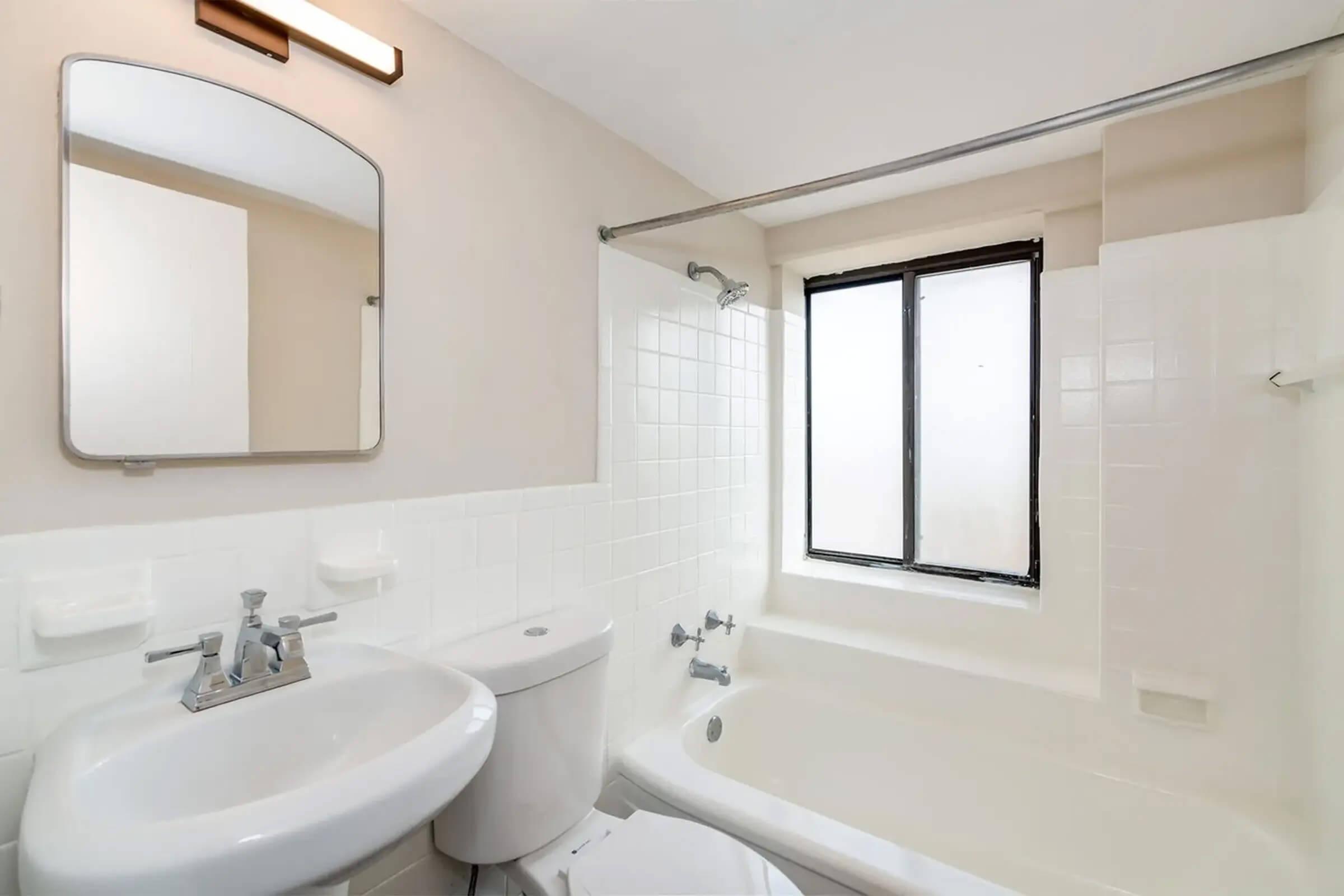 A clean and bright bathroom featuring a small tub with a shower, a white toilet, and a modern sink with a mirror above it. Natural light comes in through a window, illuminating the neutral-colored walls and tiled surfaces.