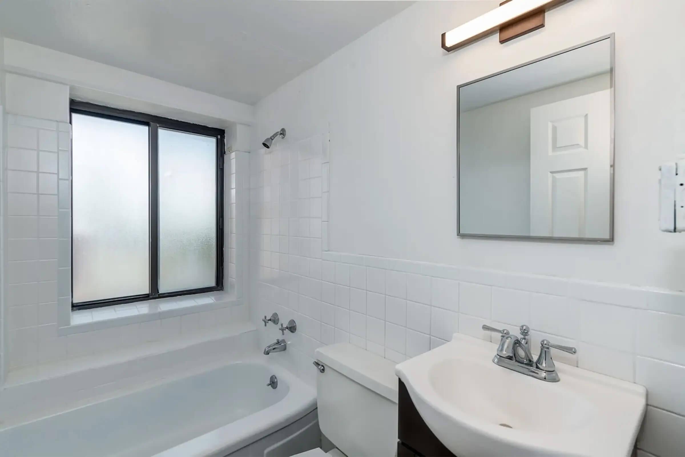 A clean, bright bathroom featuring a white bathtub, toilet, and sink. The walls are tiled in white, and there is a mirror above the sink. A window with frosted glass allows natural light while maintaining privacy. The overall aesthetic is simple and modern.