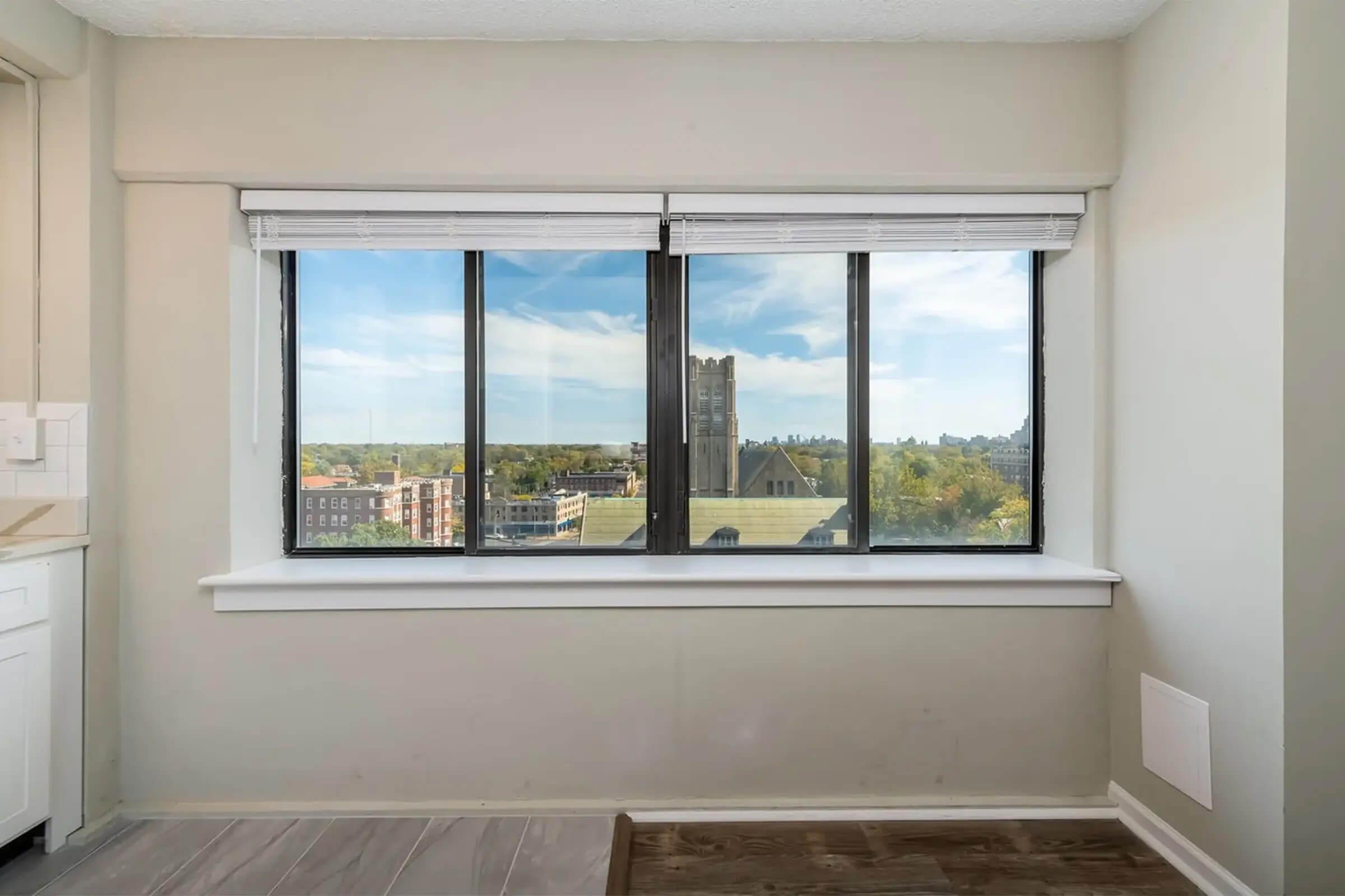 A bright room with large windows showcasing a view of the cityscape beyond, featuring buildings and greenery. The interior walls are light-colored, and the floor has a wooden finish. Sunlight illuminates the space, creating a welcoming atmosphere.