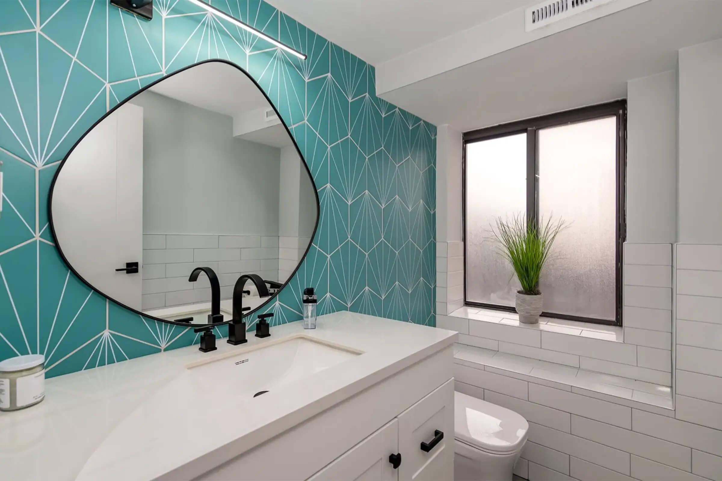 Modern bathroom featuring a geometric teal accent wall, a large oval mirror with a black frame, a white vanity with a sink, and a stylish black faucet. Natural light filters through a window with frosted glass, and a small potted plant adds a touch of greenery on the windowsill.