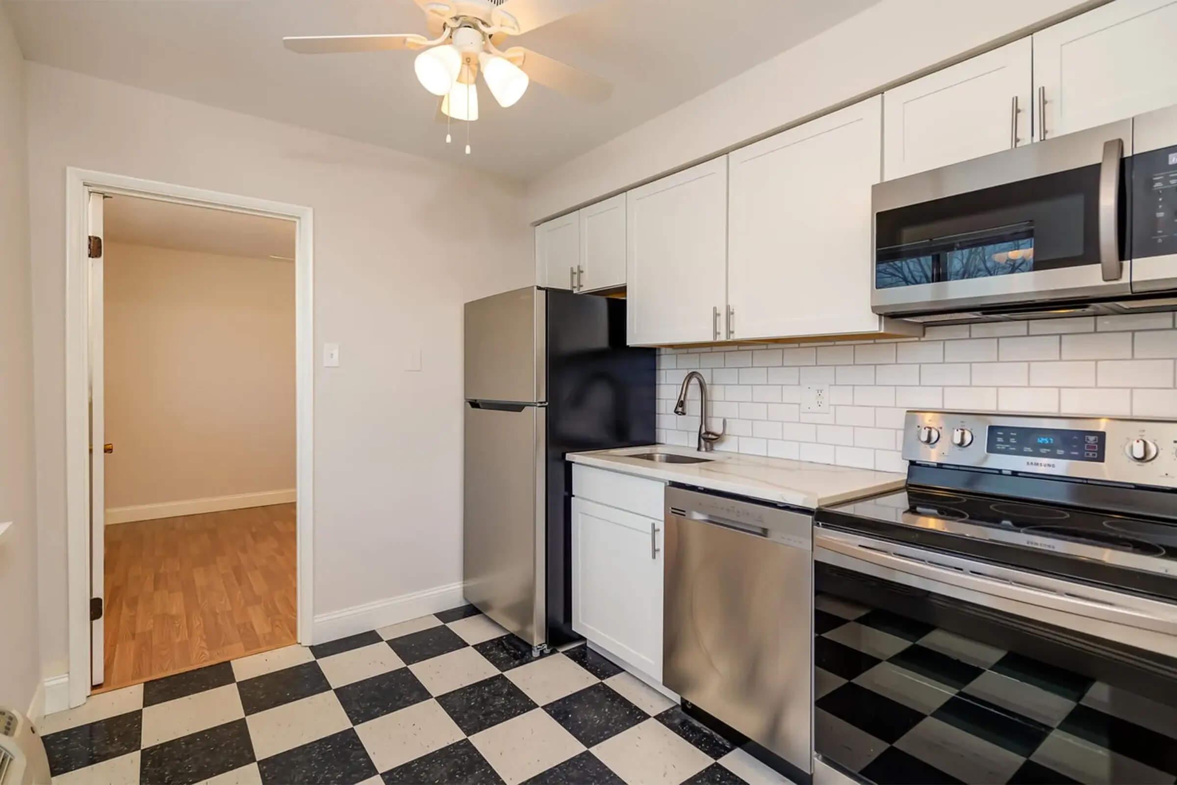 Modern kitchen featuring stainless steel appliances, including a refrigerator and oven. The cabinets are white with a sleek finish, and the countertop is light-colored. The floor has a black and white checkerboard pattern. A ceiling fan is installed above, and a doorway leads to an adjacent room.