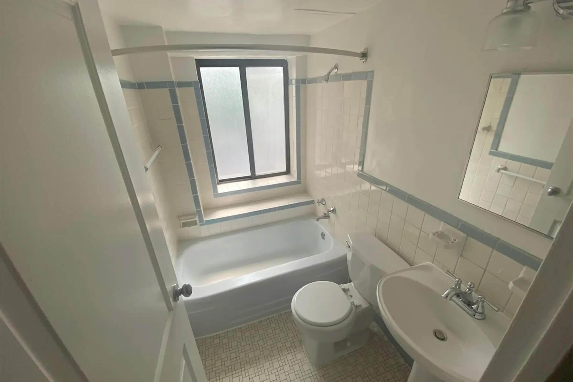 A small, well-lit bathroom featuring a bathtub with a showerhead, a toilet, and a sink. The walls are tiled in light colors with blue accents, and there’s a window allowing natural light to enter. A mirrored cabinet is mounted above the sink, and the floor is tiled with small patterned squares.