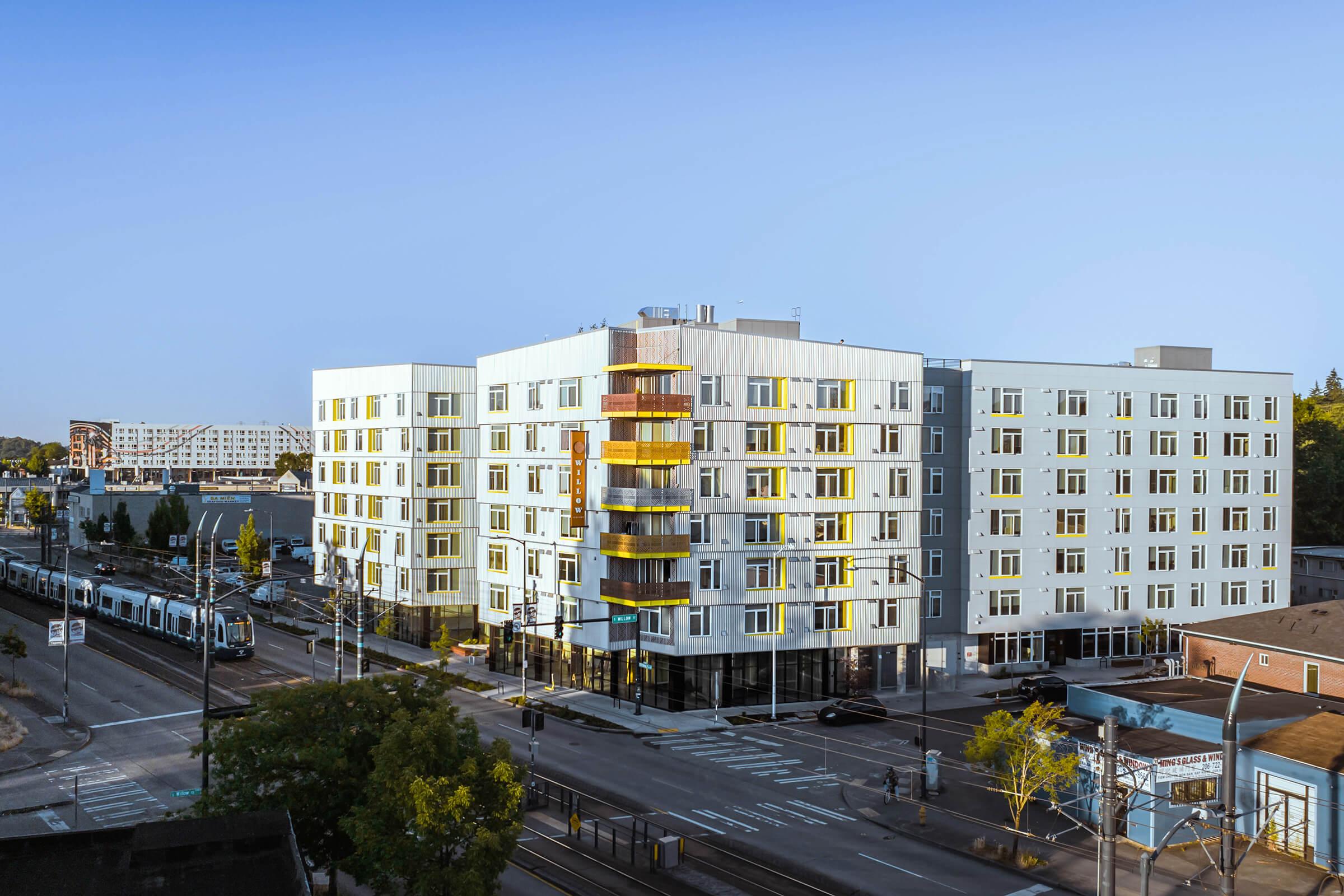 A modern multistory building featuring a combination of white and yellow exterior, with large windows and a distinctive staircase on the facade. The structure is located at a street intersection, with a light rail train and several trees visible in the foreground. Clear blue sky in the background.