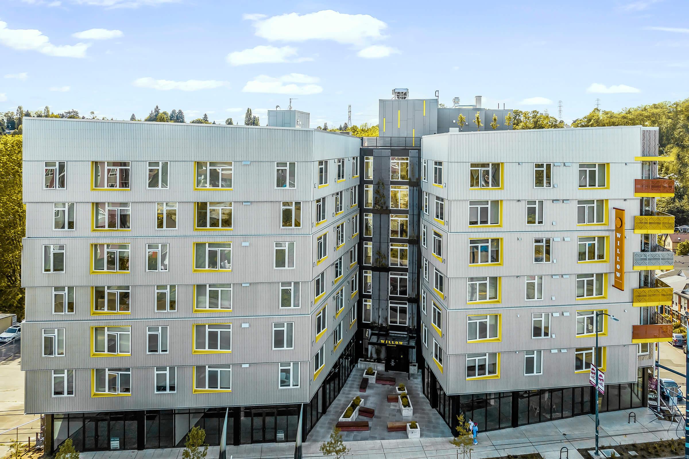 A modern five-story apartment building featuring a mix of gray and yellow exterior accents. The structure includes large windows and a landscaped area with seating in front. The building's design is contemporary, with a sleek, urban aesthetic. Trees and nearby infrastructure are visible in the background.