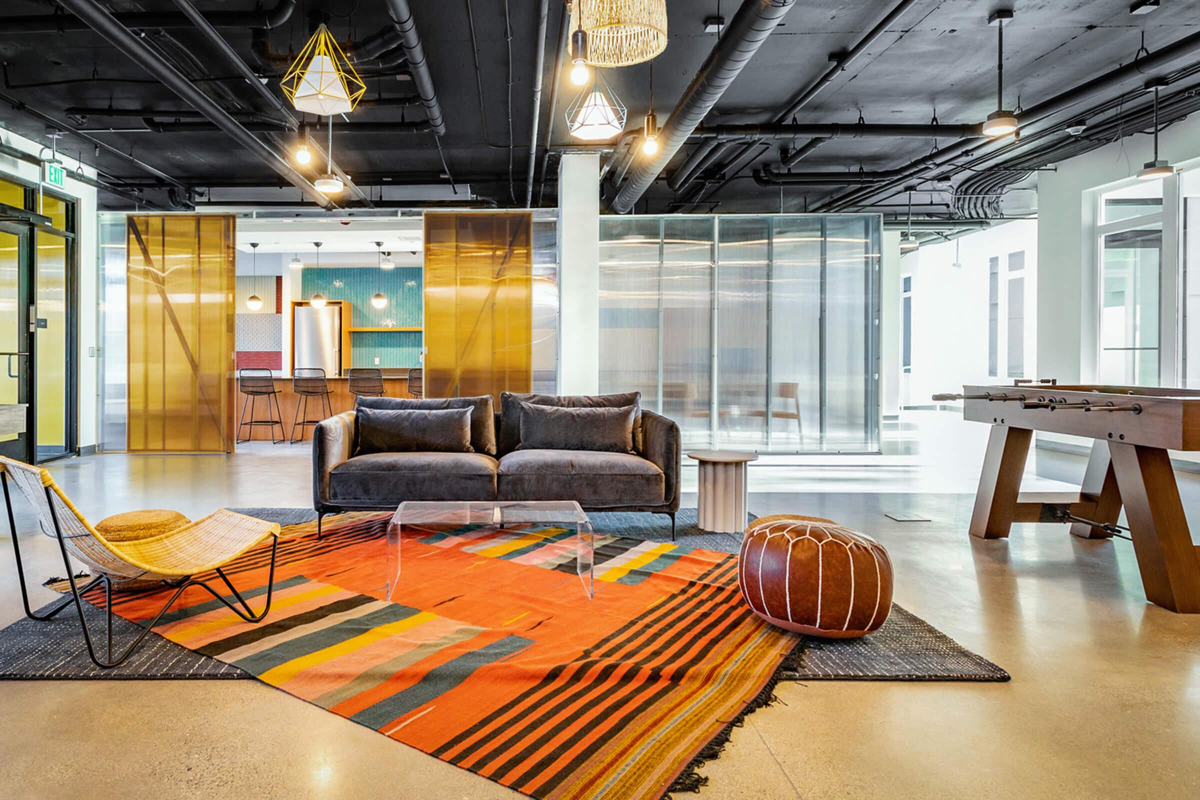 A modern lounge area featuring a stylish gray couch, a colorful area rug, and a playful accent chair. The space has large windows, industrial lighting, and an inviting atmosphere, highlighted by wooden elements and a foosball table in the background.