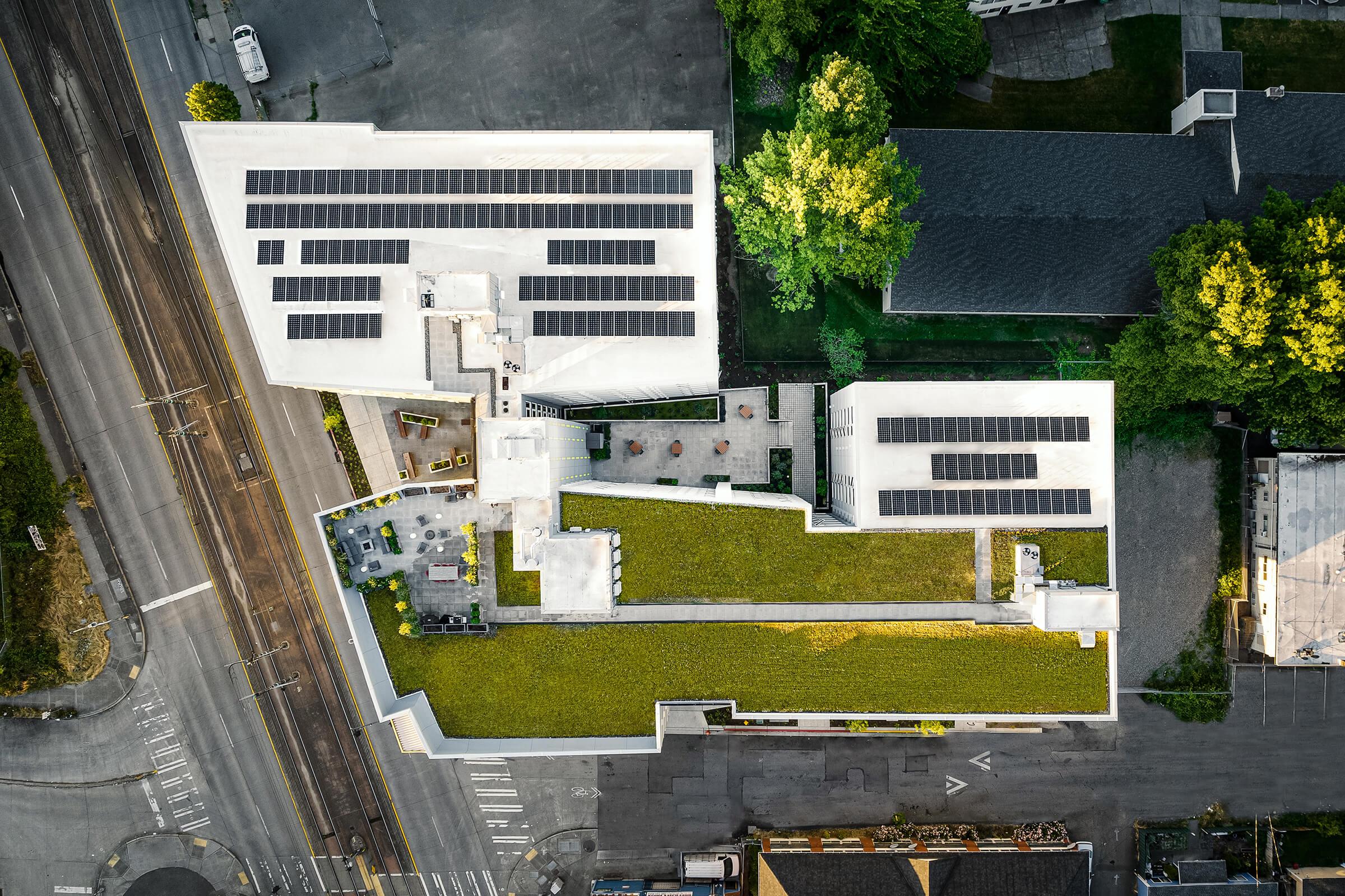 An aerial view of a modern building complex featuring multiple rooftops with solar panels. The design includes green spaces with gardens and seating areas. Surrounding roads and nearby structures can be seen, emphasizing the urban environment. The overall layout is geometric and environmentally conscious.