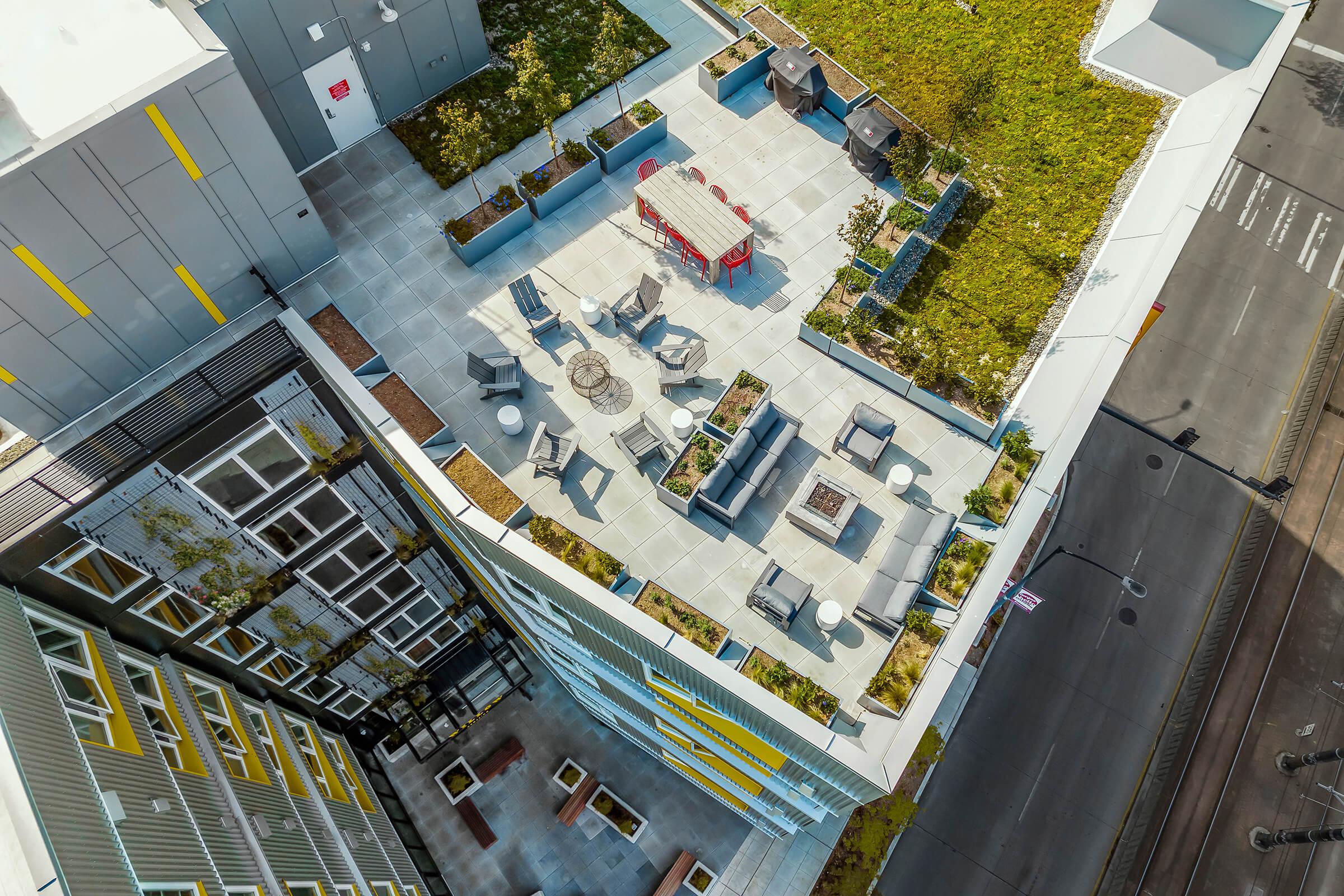 Aerial view of a modern rooftop terrace featuring seating areas, fire pits, and greenery. Brightly colored planters and furniture create an inviting space. The surrounding buildings are visible, along with a street below, providing a vibrant urban landscape.