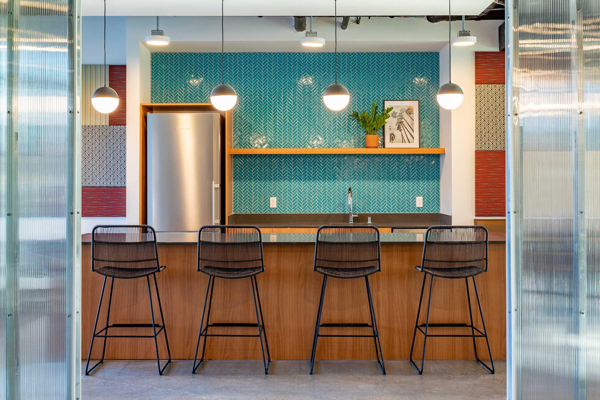 A modern kitchen featuring a sleek design with a blue patterned backsplash, wooden shelves, and bar stools. The space includes a stainless steel refrigerator, a sink, and decorative plants, illuminated by hanging pendant lights, creating a stylish and inviting atmosphere.