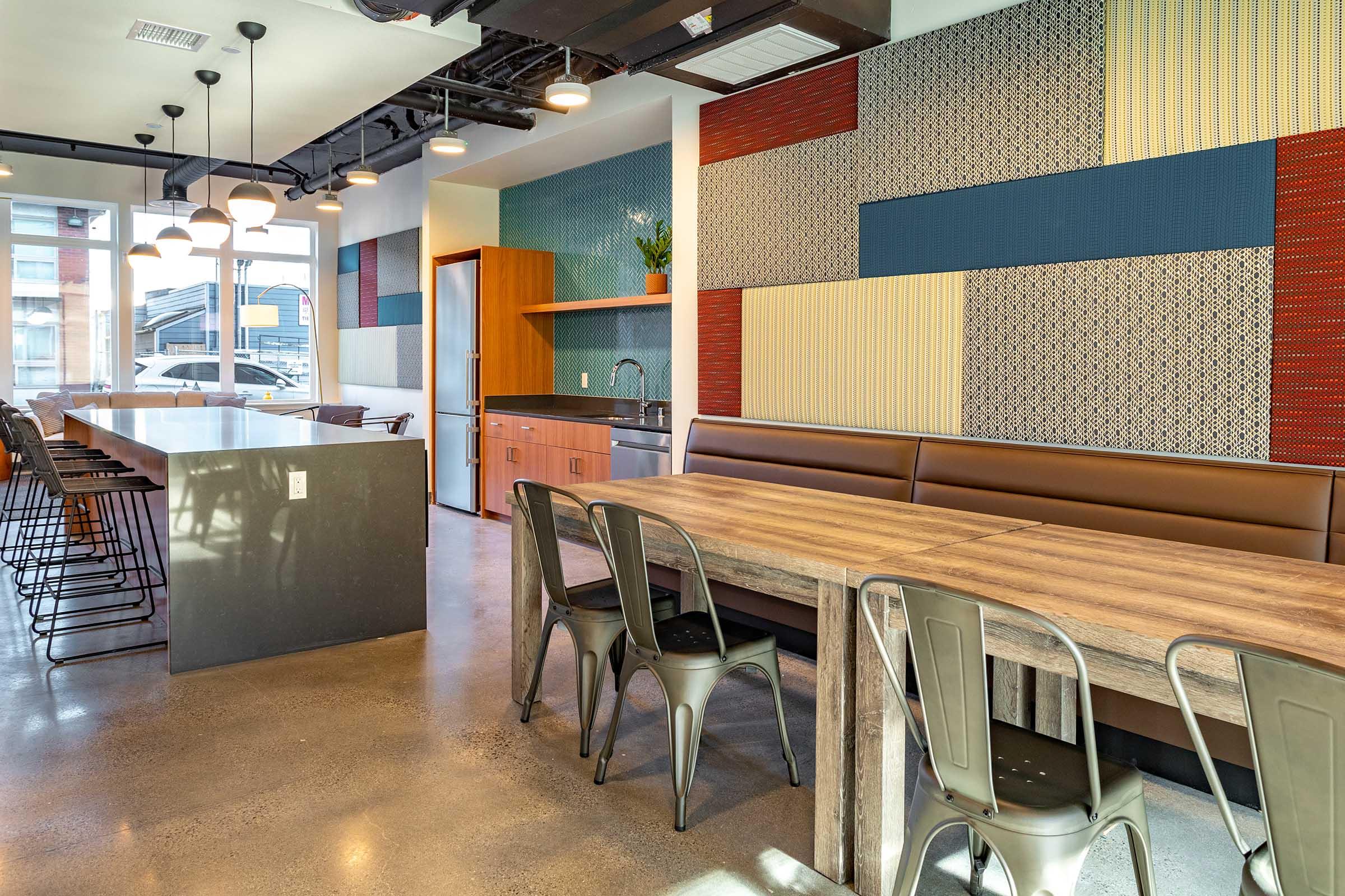 Interior of a modern dining space featuring a long wooden table with metal chairs, sleek cabinetry, and colorful textured wall panels. The area is brightly lit with pendant lights, creating a contemporary ambiance suitable for group dining or social gatherings.