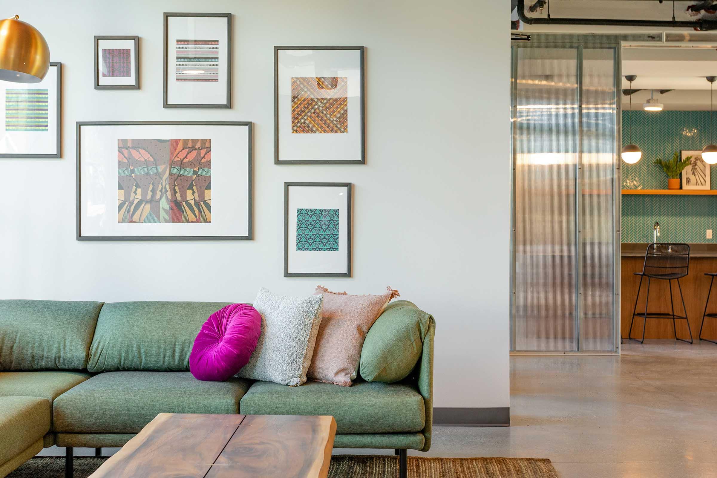 A modern living area featuring a green sofa adorned with colorful pillows, a wooden coffee table, and framed artwork on the walls. In the background, a kitchen area is visible with a bar counter and stools, illuminated by pendant lights, creating a warm and inviting atmosphere.