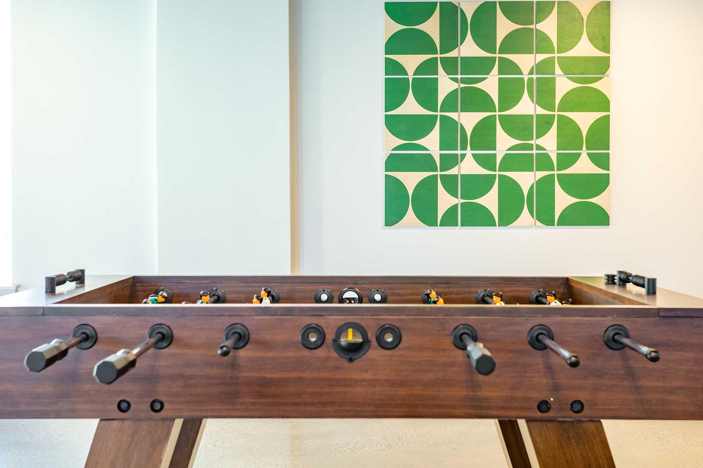 A wooden foosball table with black and gray handles in a bright room. In the background, a geometric green and white wall art piece featuring circular shapes is displayed above the table. The overall ambiance is modern and inviting.