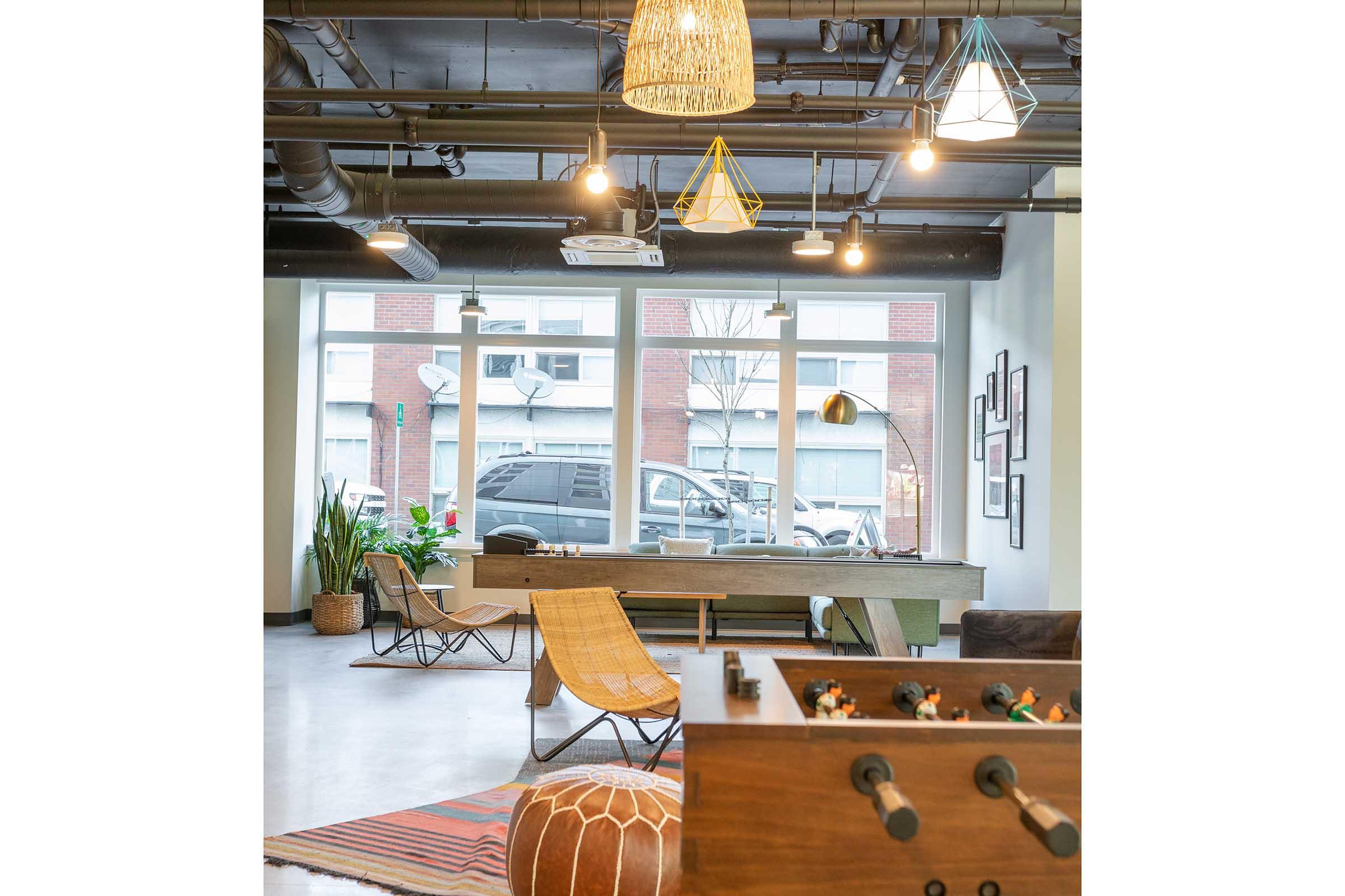 A modern interior space featuring a pool table, stylish lounge chairs, and decorative lighting. Large windows illuminate the area, showcasing a view of parked cars outside. Potted plants add a touch of greenery, while an elegant rug enhances the inviting atmosphere.