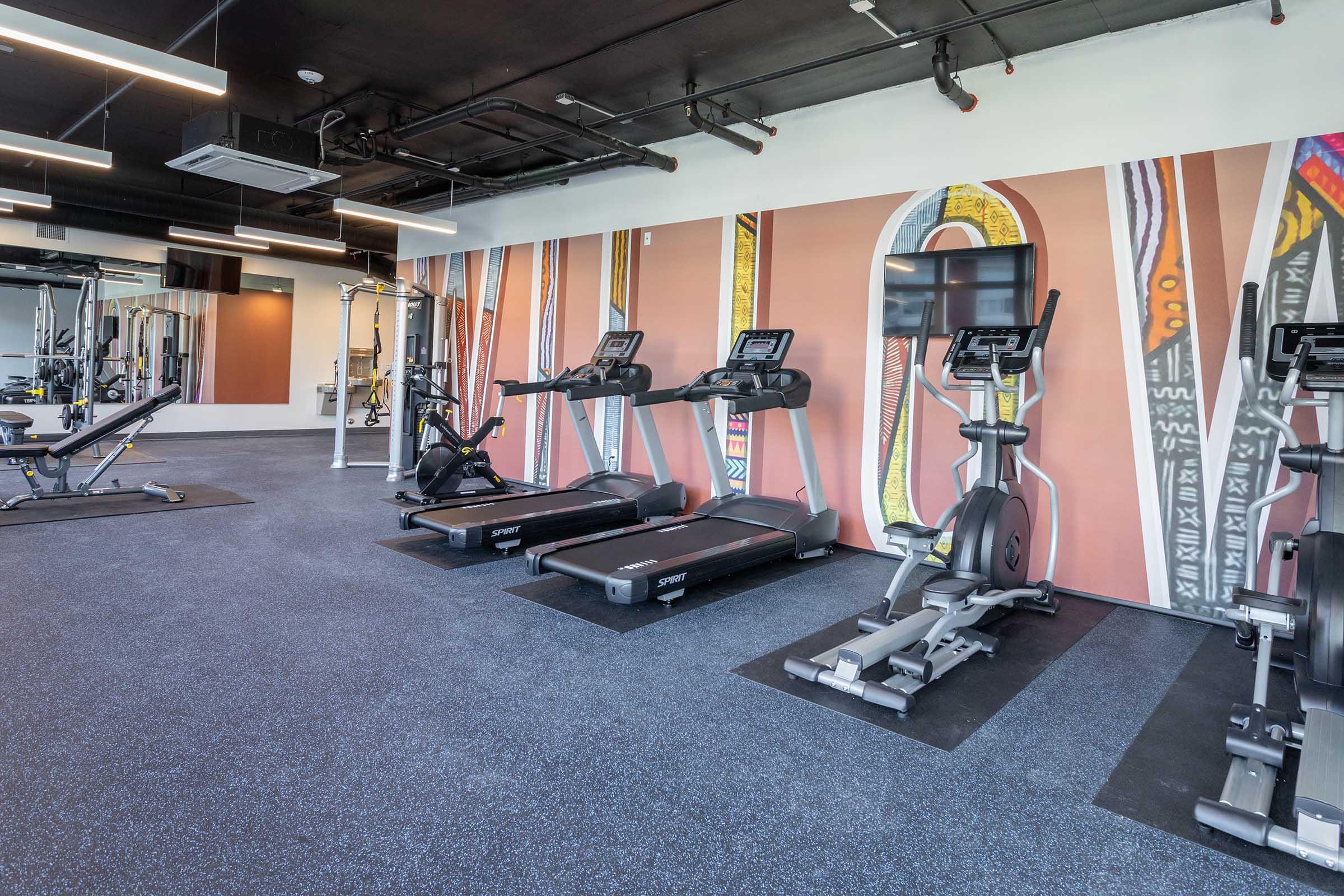 A modern gym interior featuring cardio equipment like treadmills and ellipticals, with a colorful mural on the wall. The space is well-lit and has a clean, contemporary design, highlighting various workout stations and a rubberized floor.