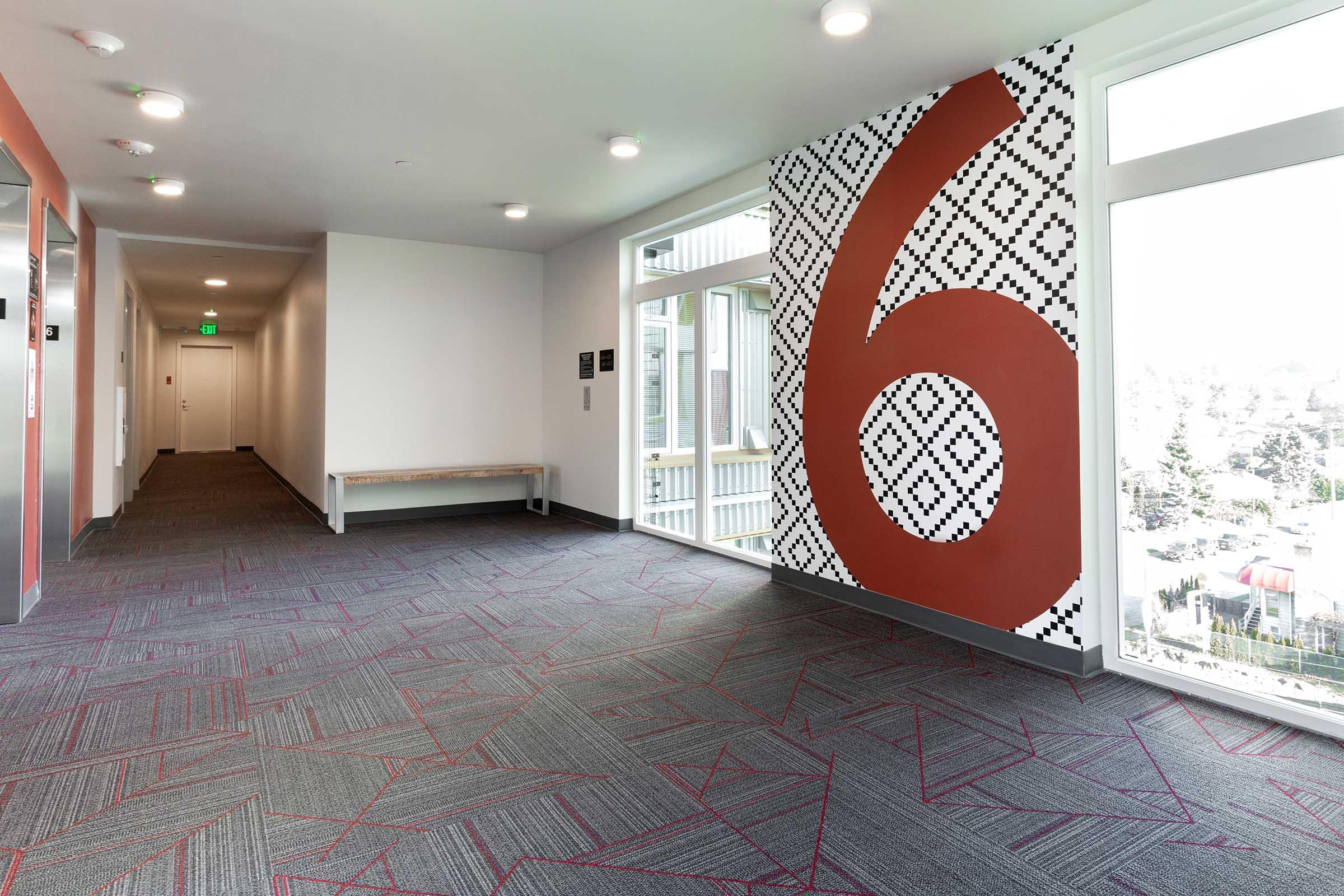 A brightly lit hallway featuring a patterned wall with the number "6" in red. There is a bench along one side and doors leading to rooms. The carpet has a geometric design, and large windows allow natural light to enter, providing a view of the outside.