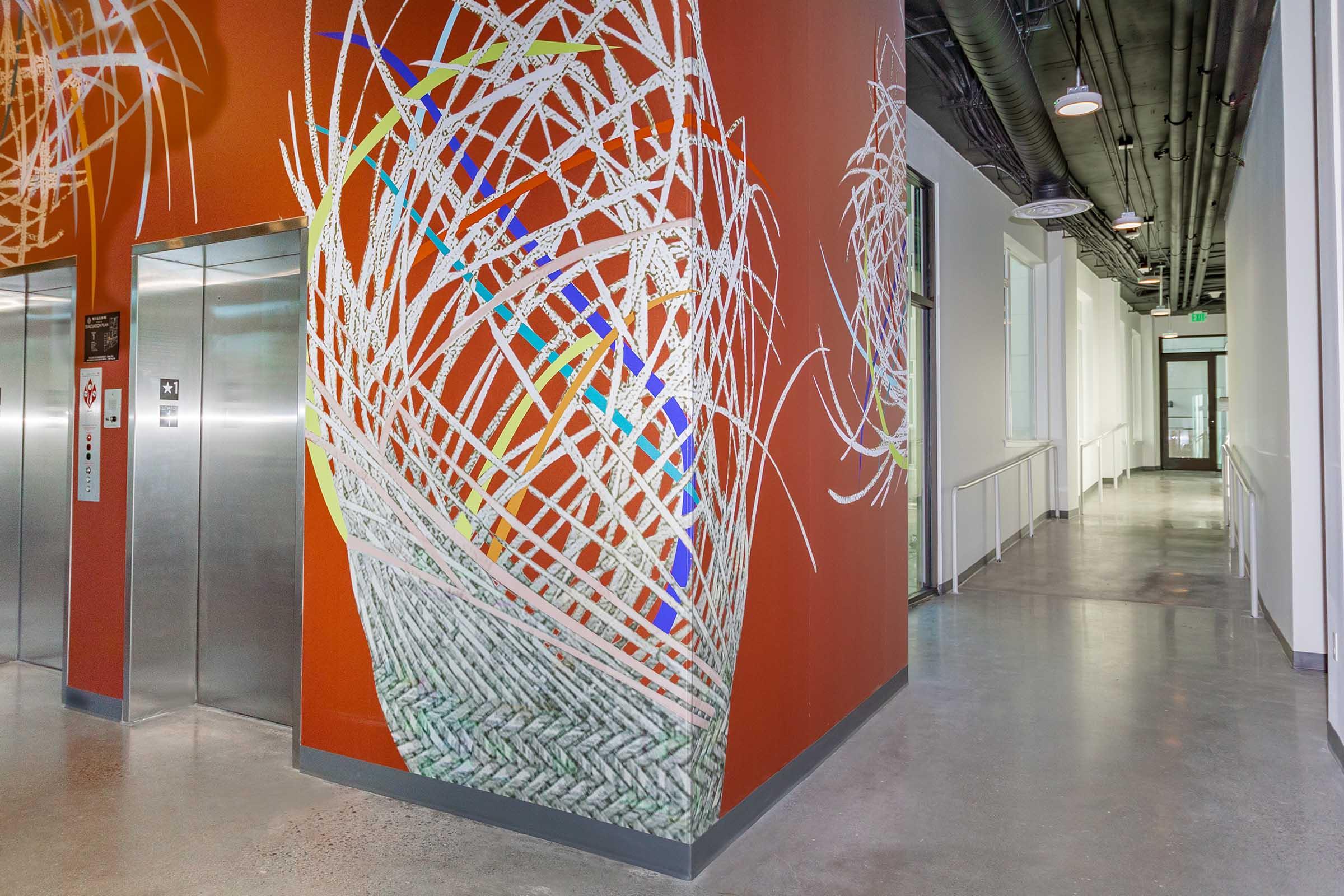 A vibrant mural decorates a red wall in a modern corridor, featuring abstract, woven designs with colorful elements. The space includes elevator doors to the left and is well-lit, with large windows along the right side, leading to a clean, polished floor.