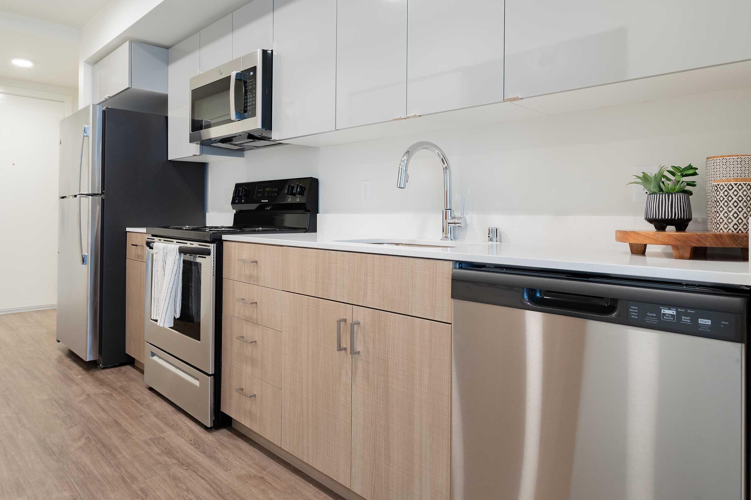 Modern kitchen featuring a stainless steel refrigerator, microwave, and oven. Light wood cabinets contrast with sleek gray countertops. A stainless steel dishwasher is integrated beneath the counter. A small plant and decorative container are displayed on a wooden shelf, adding a touch of greenery to the space.