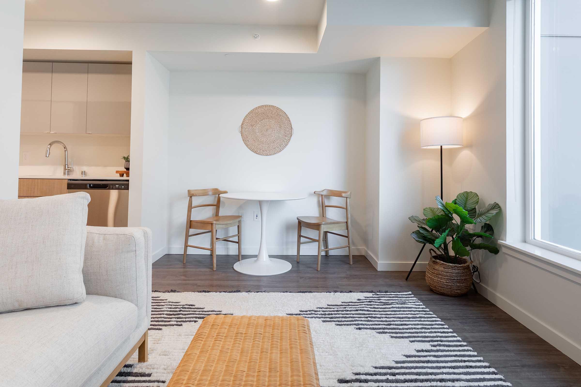 A cozy interior space featuring a small round table with two wooden chairs, a light-colored sofa, and a decorative rug. A large window lets in natural light, accompanied by a standing lamp and a potted plant in a woven basket, creating a warm and inviting atmosphere.