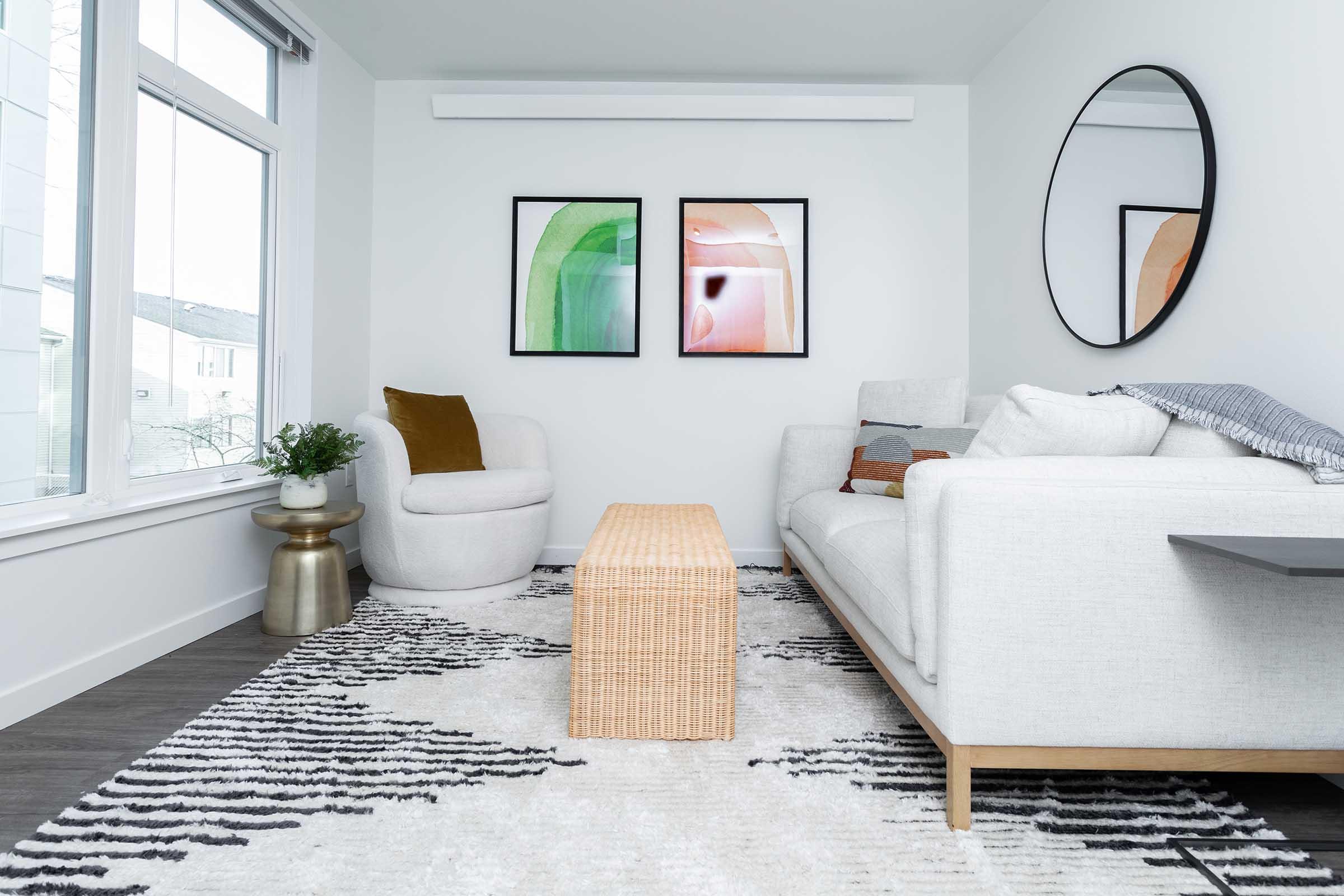 A modern living room featuring a light-colored sofa, a round chair, and a woven coffee table on a patterned rug. Two abstract framed artworks hang on the wall, and a circular mirror reflects the space. Natural light streams in through large windows, creating a bright and airy atmosphere.