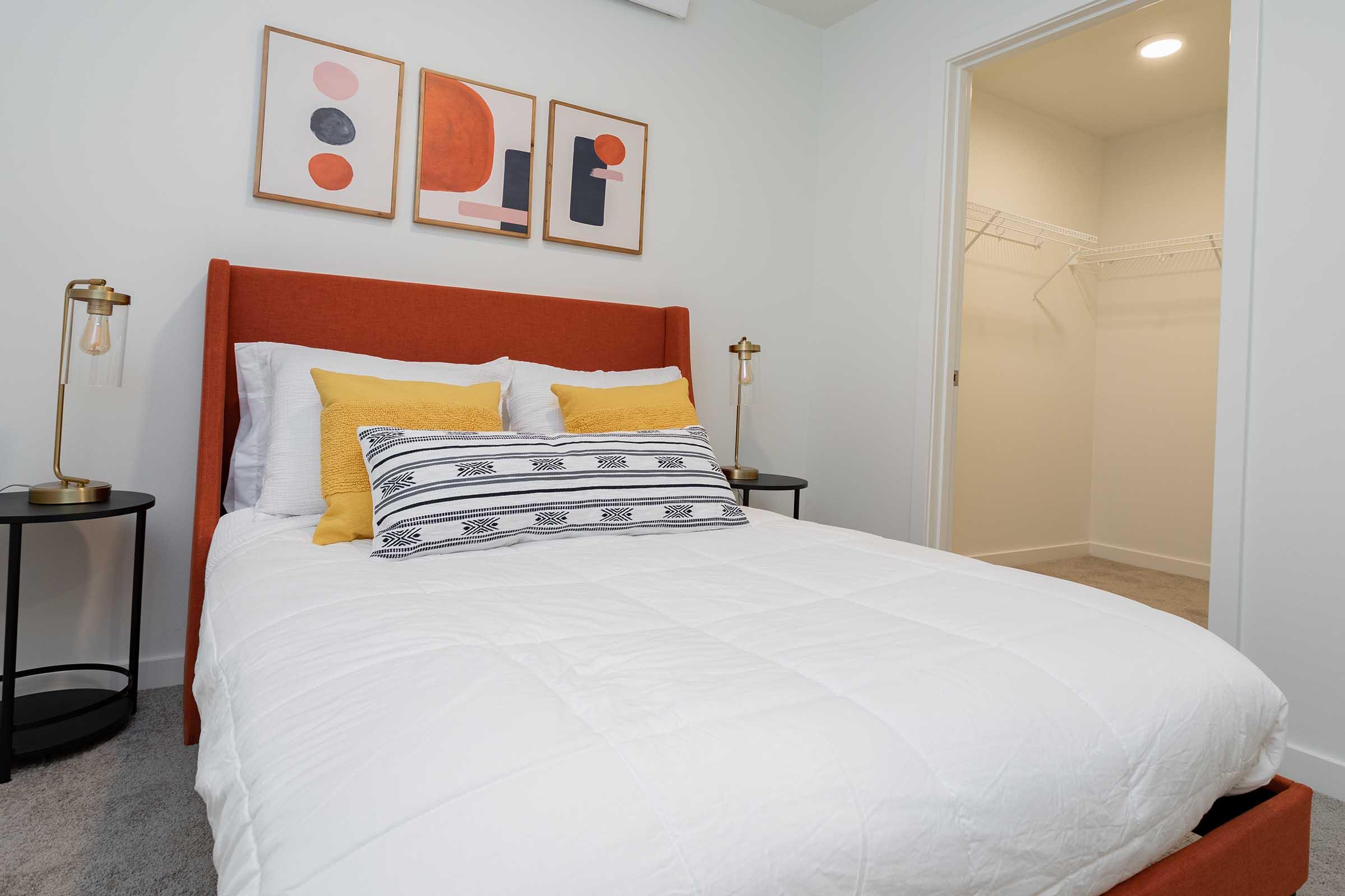 A modern bedroom featuring a stylish orange bed frame with white bedding and decorative yellow pillows. Above the bed are three abstract art pieces. Two bedside tables with lamps are on either side of the bed. In the background, there's a closet with open shelving. The walls are light-colored and the carpet is neutral.