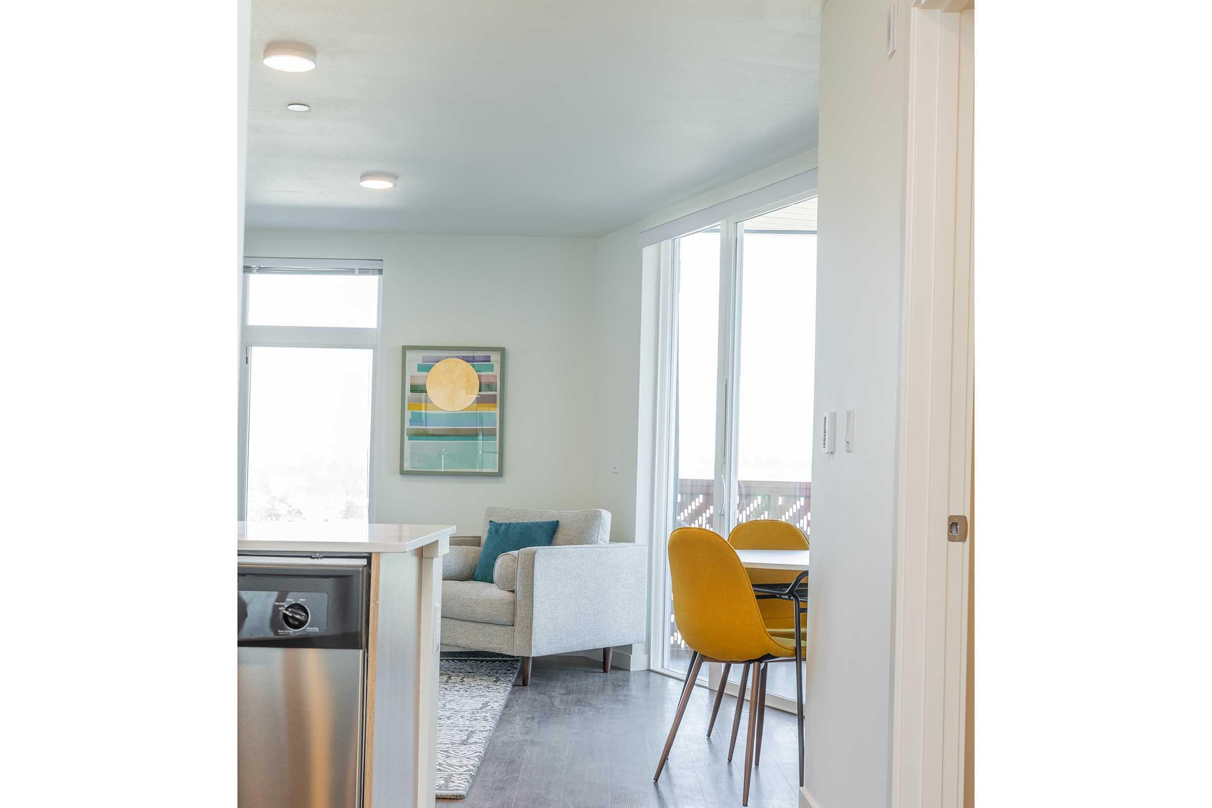 A bright, modern living space with an open layout. The image shows a cozy seating area with a gray armchair and a decorative pillow, a dining table with yellow chairs, and large windows providing natural light. Artwork hangs on the wall, and the floor features a light-colored rug.