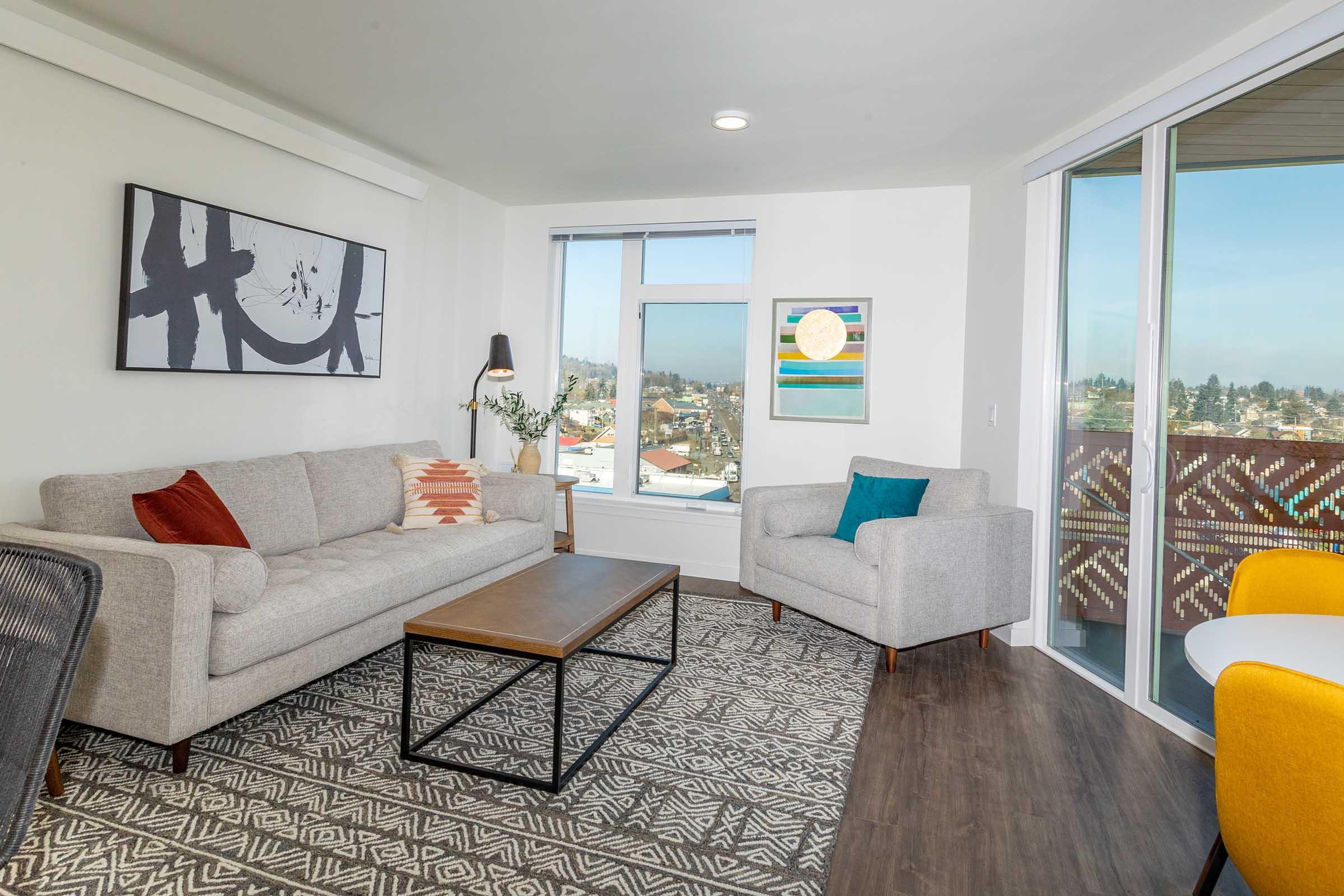 A modern living room with a light gray sofa, a light gray armchair, and a wooden coffee table on a patterned rug. There are large windows offering a view of the outdoors, and wall decor including abstract art and a circular piece. The space is bright and inviting with a cozy atmosphere.