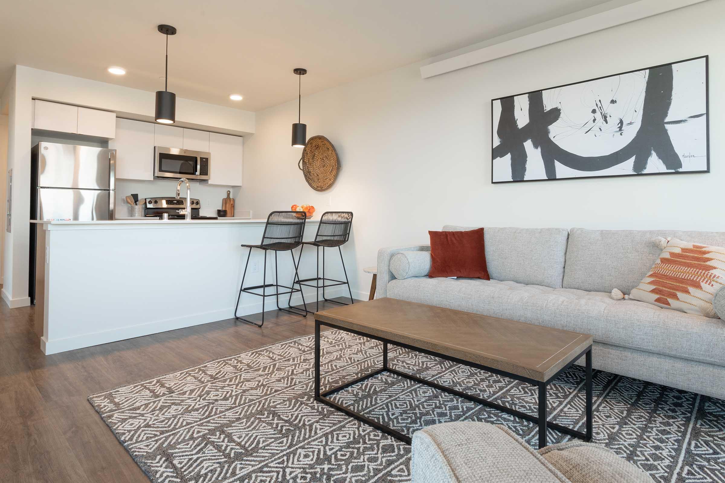 A modern living space featuring a light gray sofa with decorative pillows, a wooden coffee table on a patterned rug, and a minimalist kitchen area with stainless steel appliances. Two black bar stools are positioned by a kitchen counter. Artwork is displayed on the wall, adding a touch of style to the space.