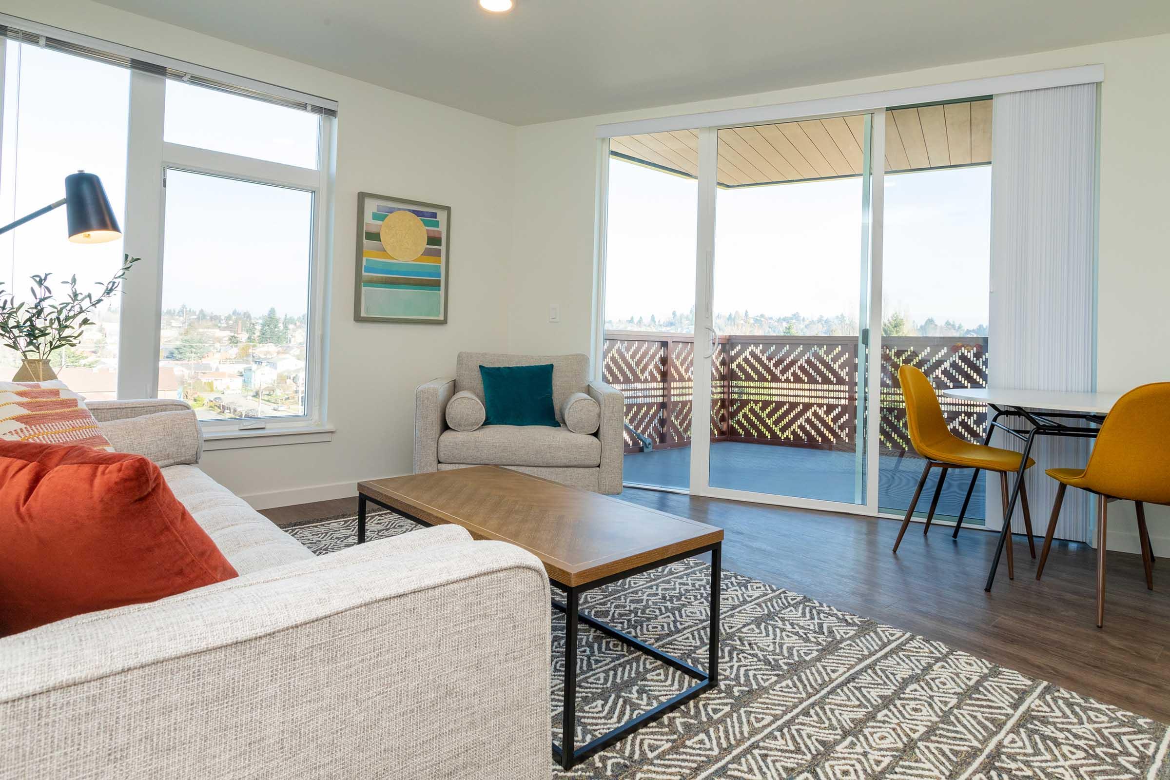 A modern living room featuring a light gray sofa with orange and patterned pillows, a wooden coffee table, and a cozy armchair. Large windows provide natural light and a view of a balcony with outdoor seating. The room has a neutral color palette and decorative wall art.