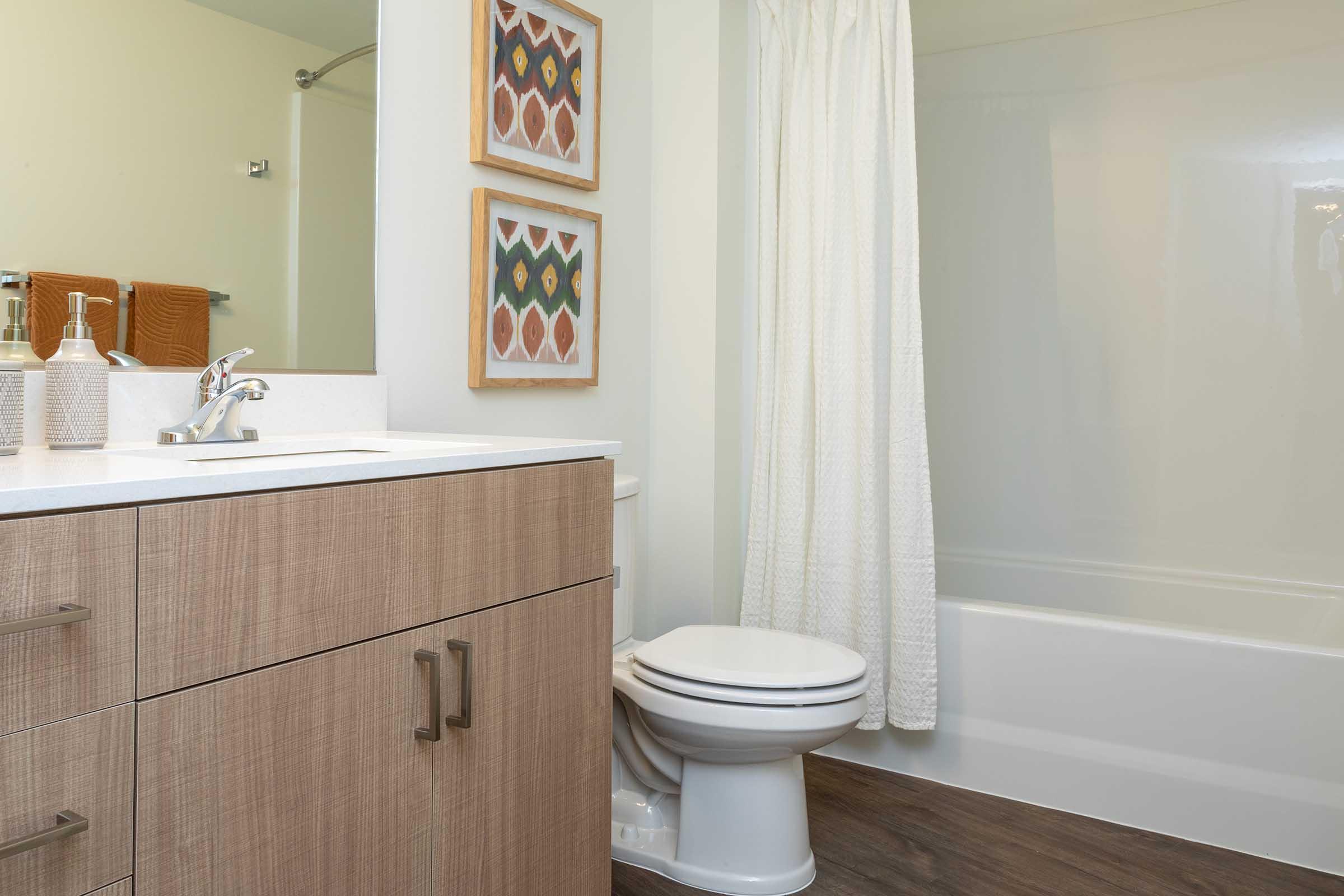 A modern bathroom featuring a white bathtub, a toilet, and a wooden vanity with a sink. The walls are painted light green, and there are two framed decorative artworks above the vanity. A white shower curtain hangs in front of the tub, and the flooring is dark wood. Towels and toiletries are neatly arranged.