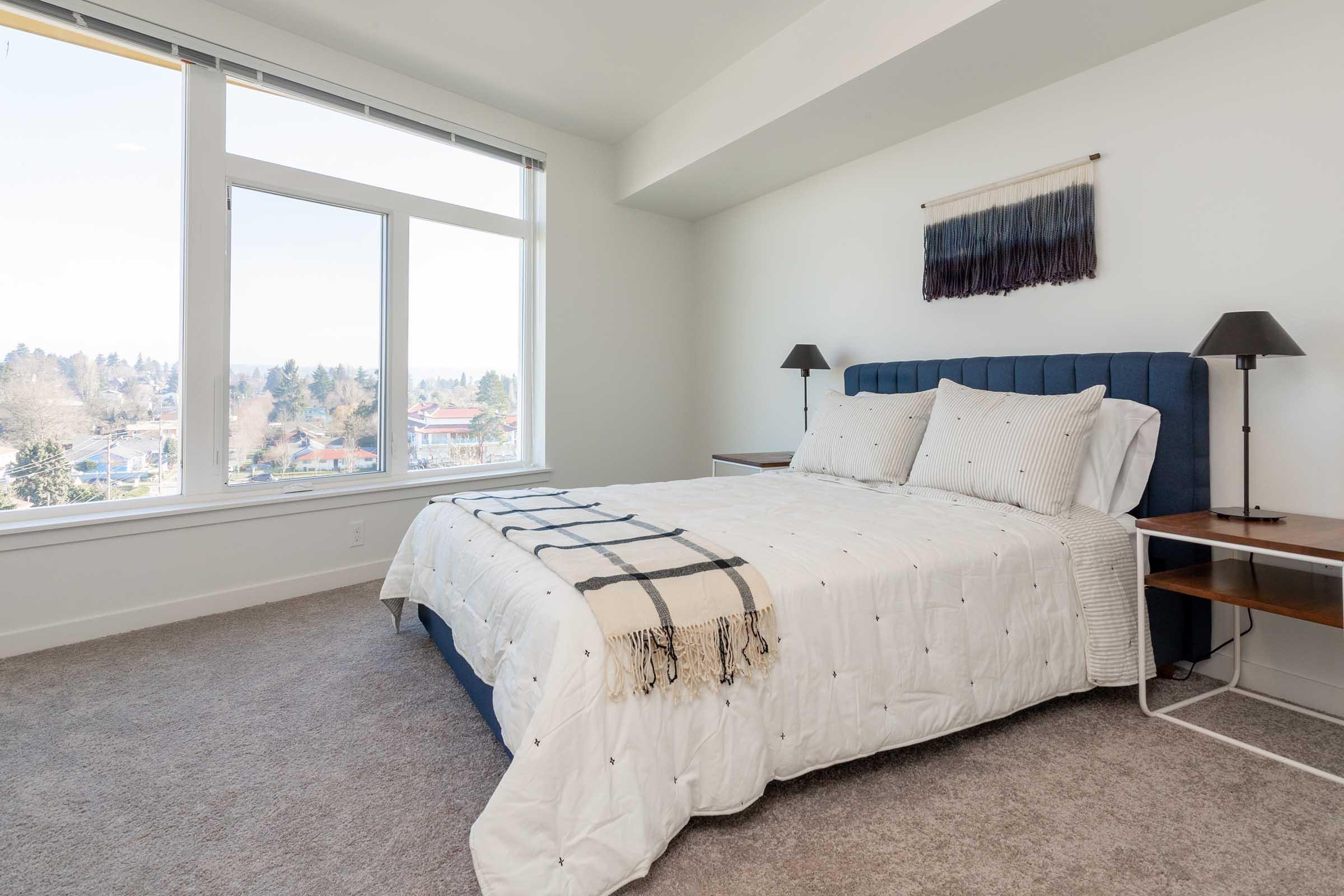 A bright and modern bedroom featuring a king-sized bed with a white comforter and decorative throw. Flanking the bed are two stylish bedside tables with lamps. A large window offers a view of the neighborhood, filling the space with natural light. The walls are painted white, and the carpet is light gray.