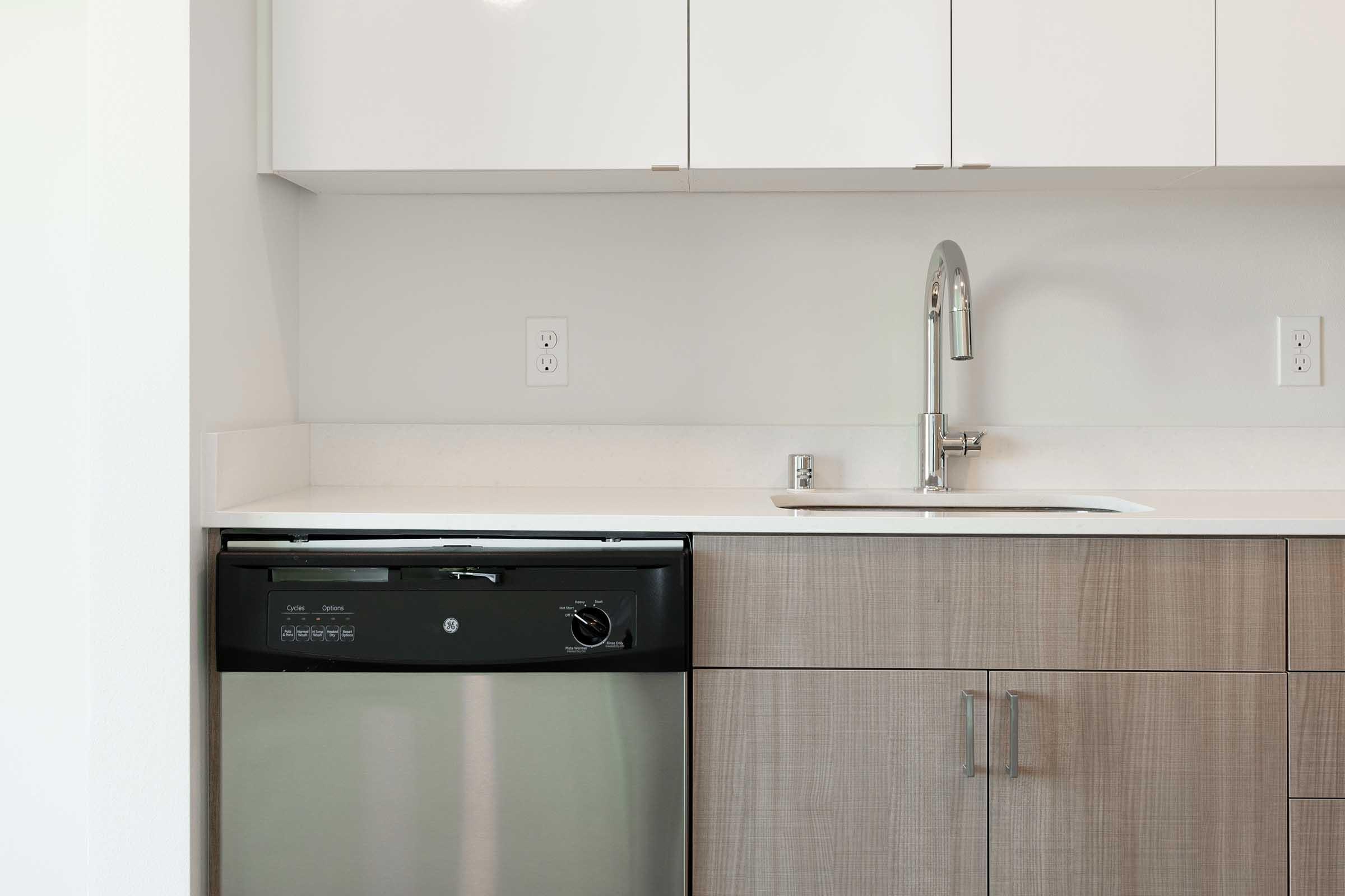 A modern kitchen featuring a stainless steel dishwasher under a countertop, paired with a sleek silver sink and a single handle faucet. The cabinets above are white, while the lower cabinets have a light wood finish, creating a clean and contemporary look.