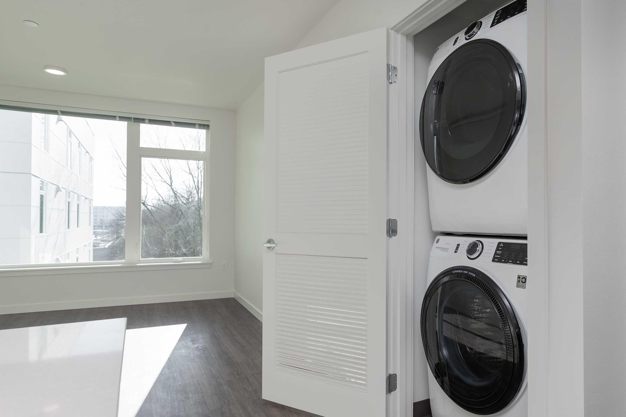 A modern laundry area featuring a stacked washer and dryer unit inside a doorway, with large windows allowing natural light to enter the space. The flooring is sleek and contemporary, and the walls are painted in neutral colors, creating a clean and airy atmosphere.