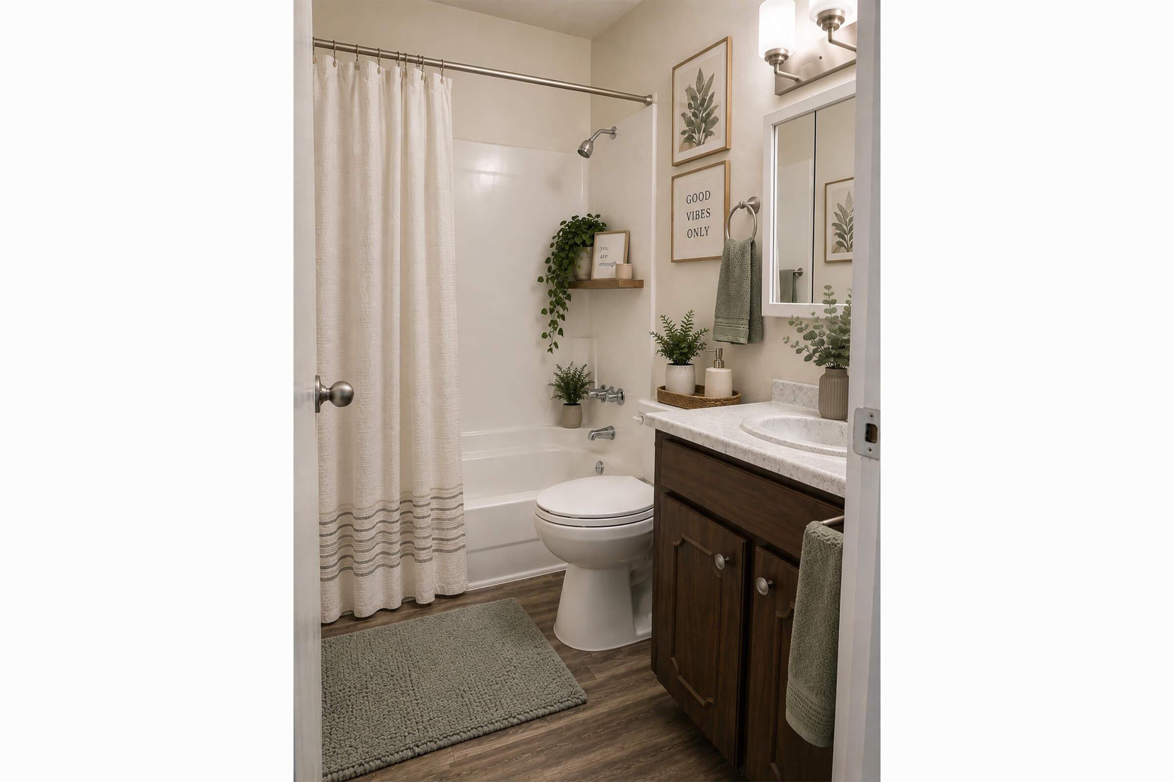 A clean, modern bathroom featuring a white bathtub with a shower curtain, a toilet, and a wooden vanity with a sink. The space is decorated with potted plants, framed artwork on the walls, and a soft rug on the floor, creating a fresh and inviting atmosphere.