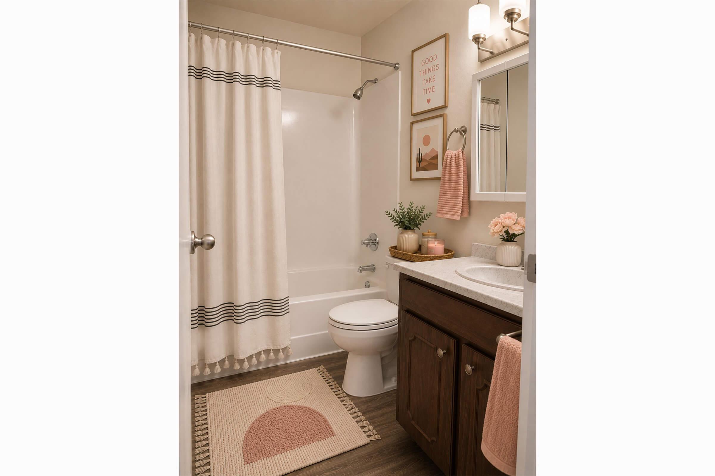 A well-decorated bathroom featuring a bathtub with a white shower curtain, a wooden vanity with a sink, a round mirror, and a decorative wall art piece. The space includes a rug with a circular design, a small plant, and fresh flowers in a vase, creating a cozy and inviting atmosphere.