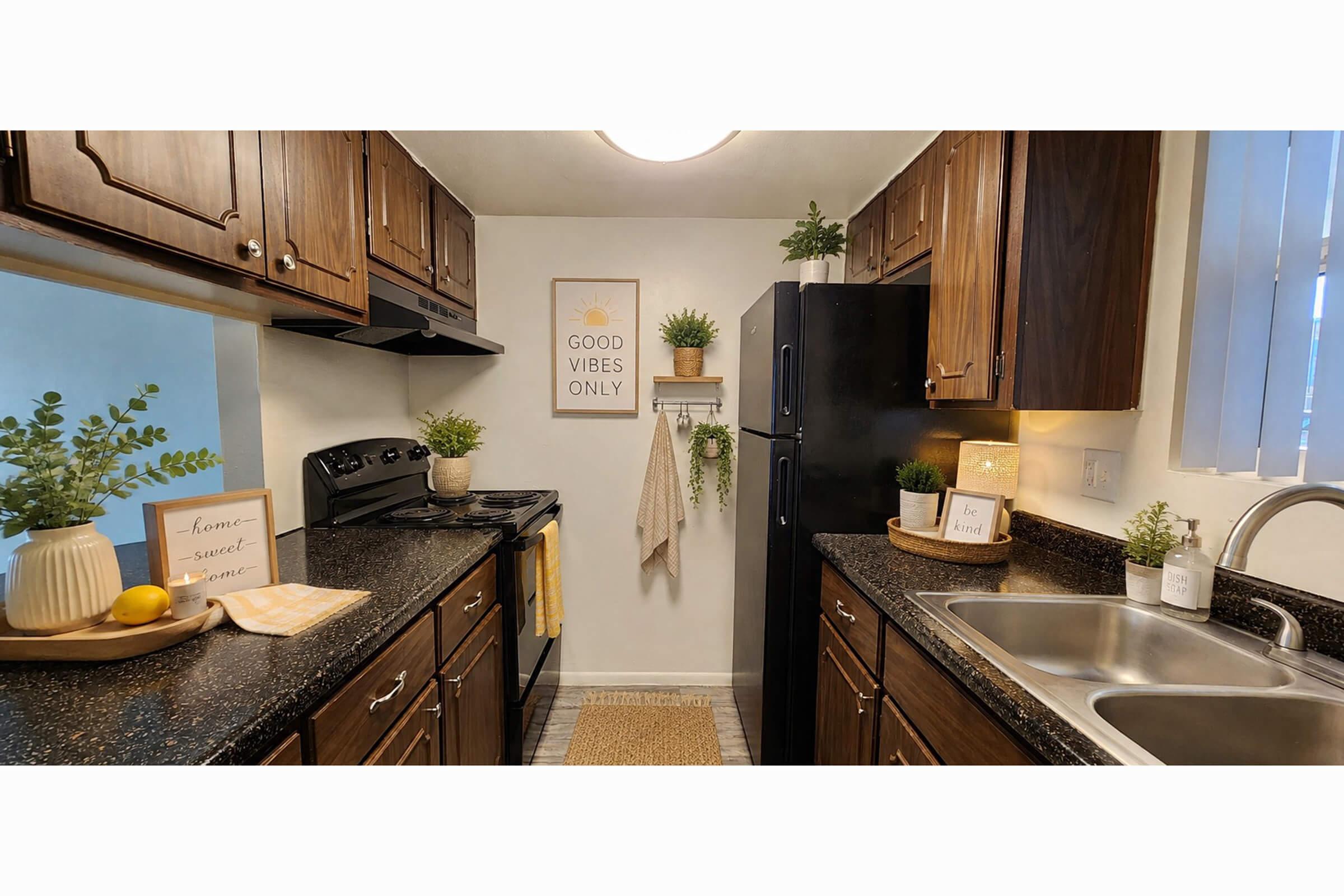 A cozy kitchen featuring dark wood cabinetry, black appliances, and a granite countertop. There are decorative plants, a "Good Vibes Only" poster, and a sign reading "home sweet home." The space is well-lit and organized, with a dish towel hanging and a rug on the floor.