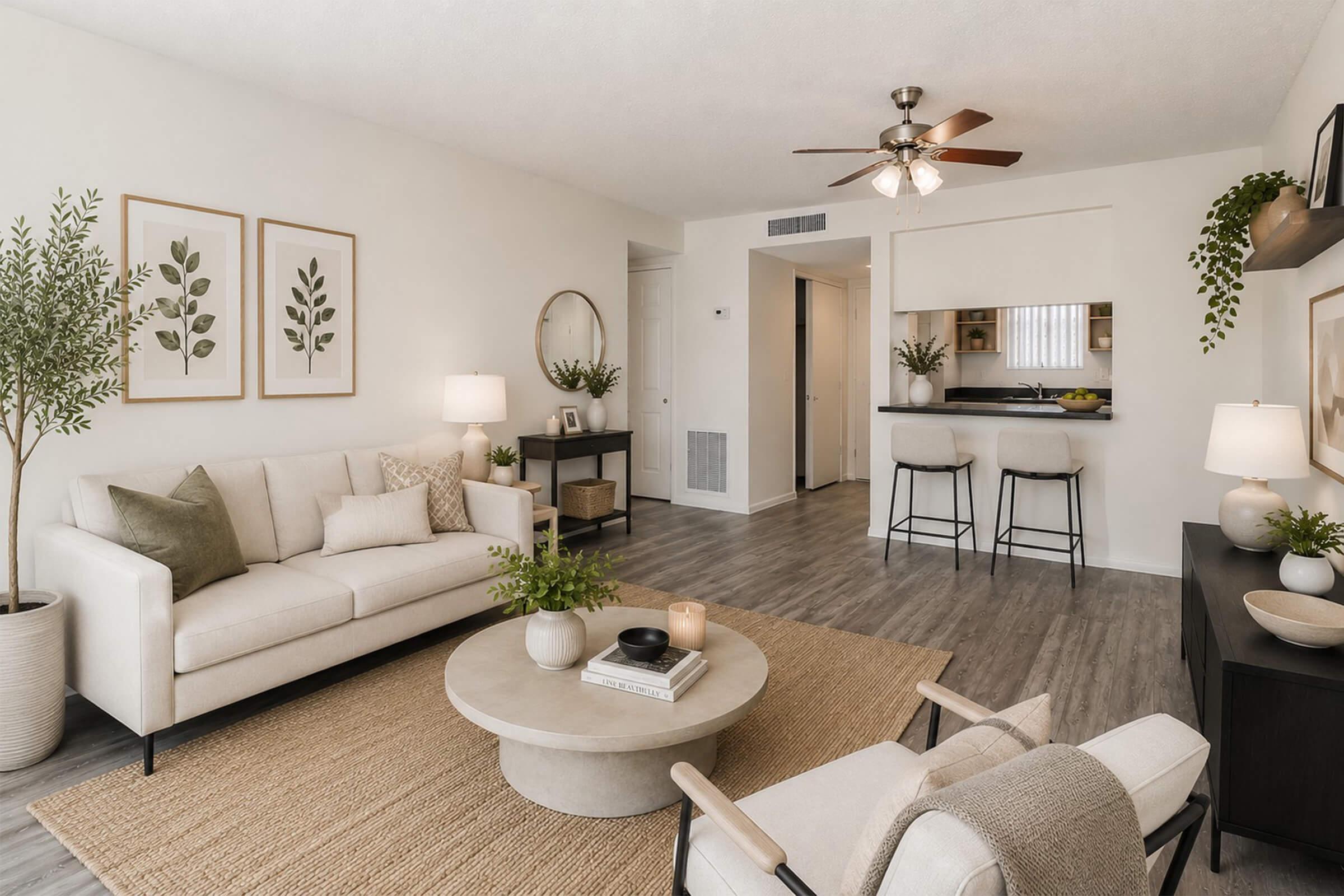 A stylish living room featuring a beige sofa with decorative pillows, a round coffee table placed on a textured rug, and two comfortable armchairs. Modern wall art with plant designs adorns the walls. A ceiling fan is visible, along with a small bar area in the background. The space is well-lit, showcasing a clean and inviting atmosphere.