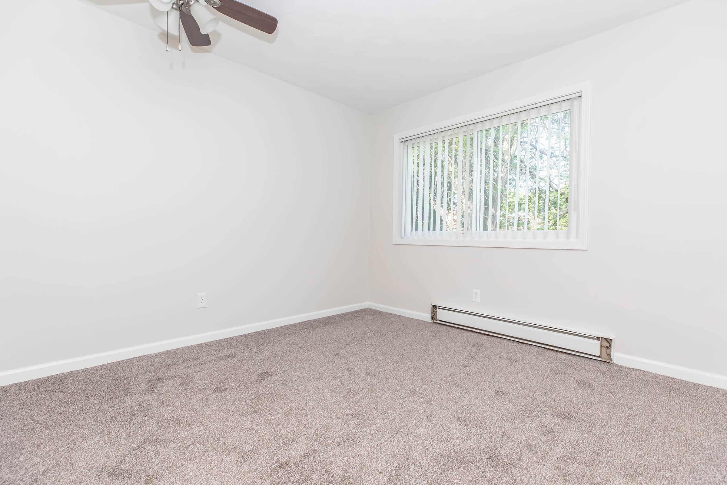 Empty room with beige walls and light brown carpet. A large window with white blinds allows natural light to enter, and a ceiling fan is visible. There is a baseboard heater beneath the window. The space appears clean and ready for furnishing, with no decor or personal items present.