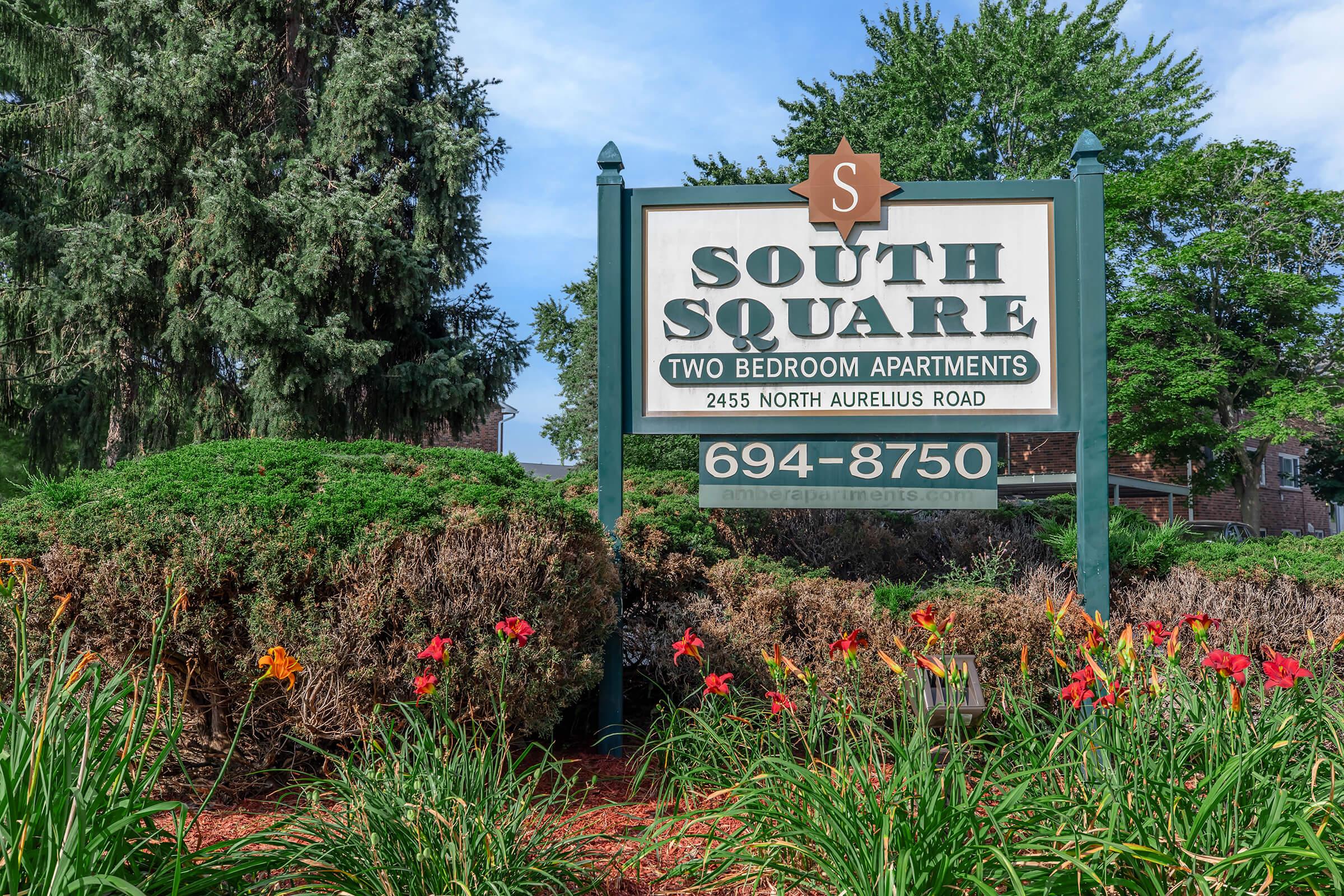 Sign for South Square Apartments featuring the text "South Square" and "Two Bedroom Apartments," along with the address "2455 North Aurelius Road" and phone number "694-8750." Surrounding the sign is lush greenery and colorful flowers.