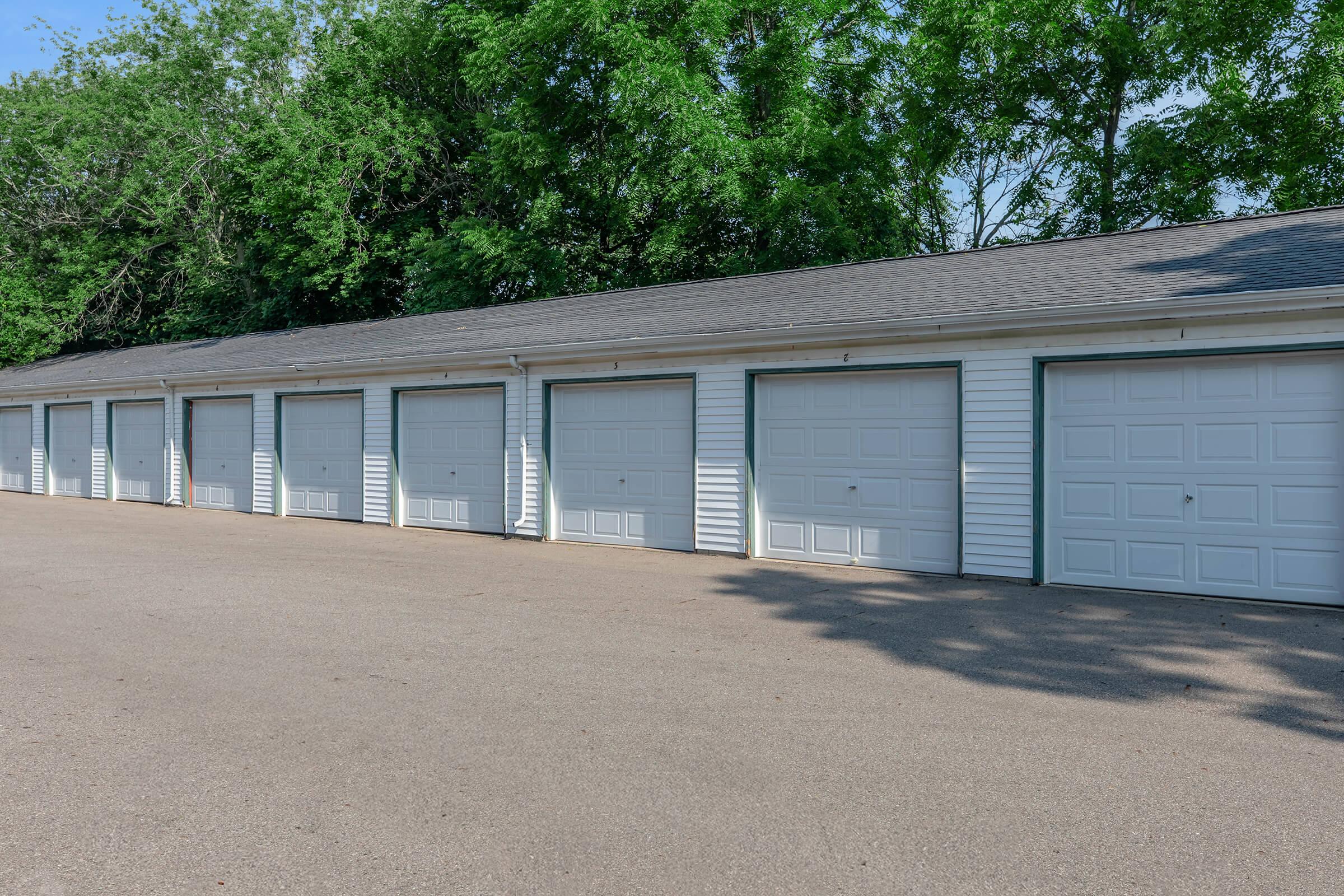 Row of closed, white garage doors lined along a paved driveway, shaded by trees in the background. The garages have green trim and are situated in a well-kept area. The scene is sunny, highlighting the neatness of the surroundings.
