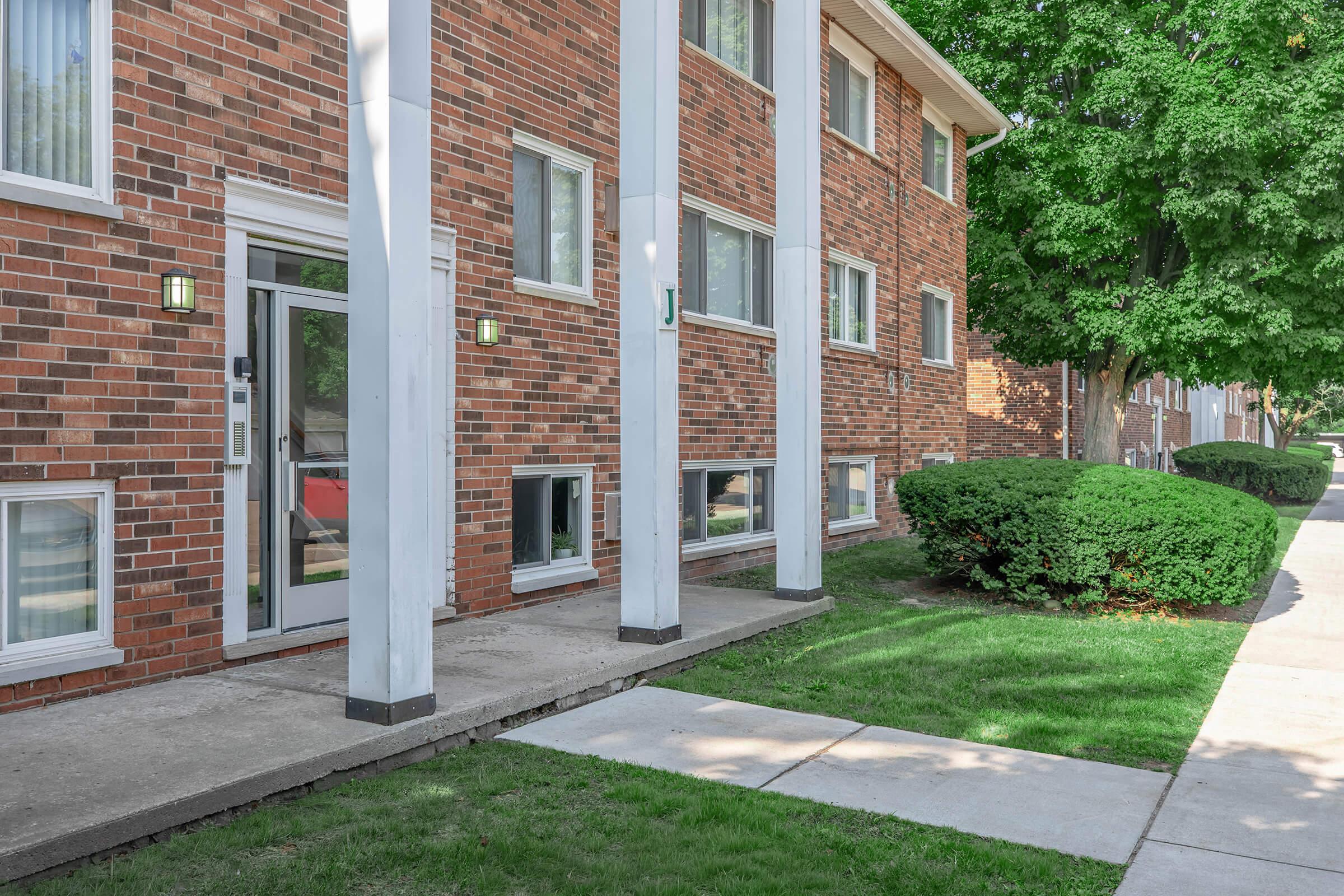 Exterior view of a brick apartment building featuring large windows and white columns. A concrete walkway leads to the entrance door, which is flanked by two wall-mounted lights. The landscaping includes well-trimmed bushes and a grassy area alongside the sidewalk.