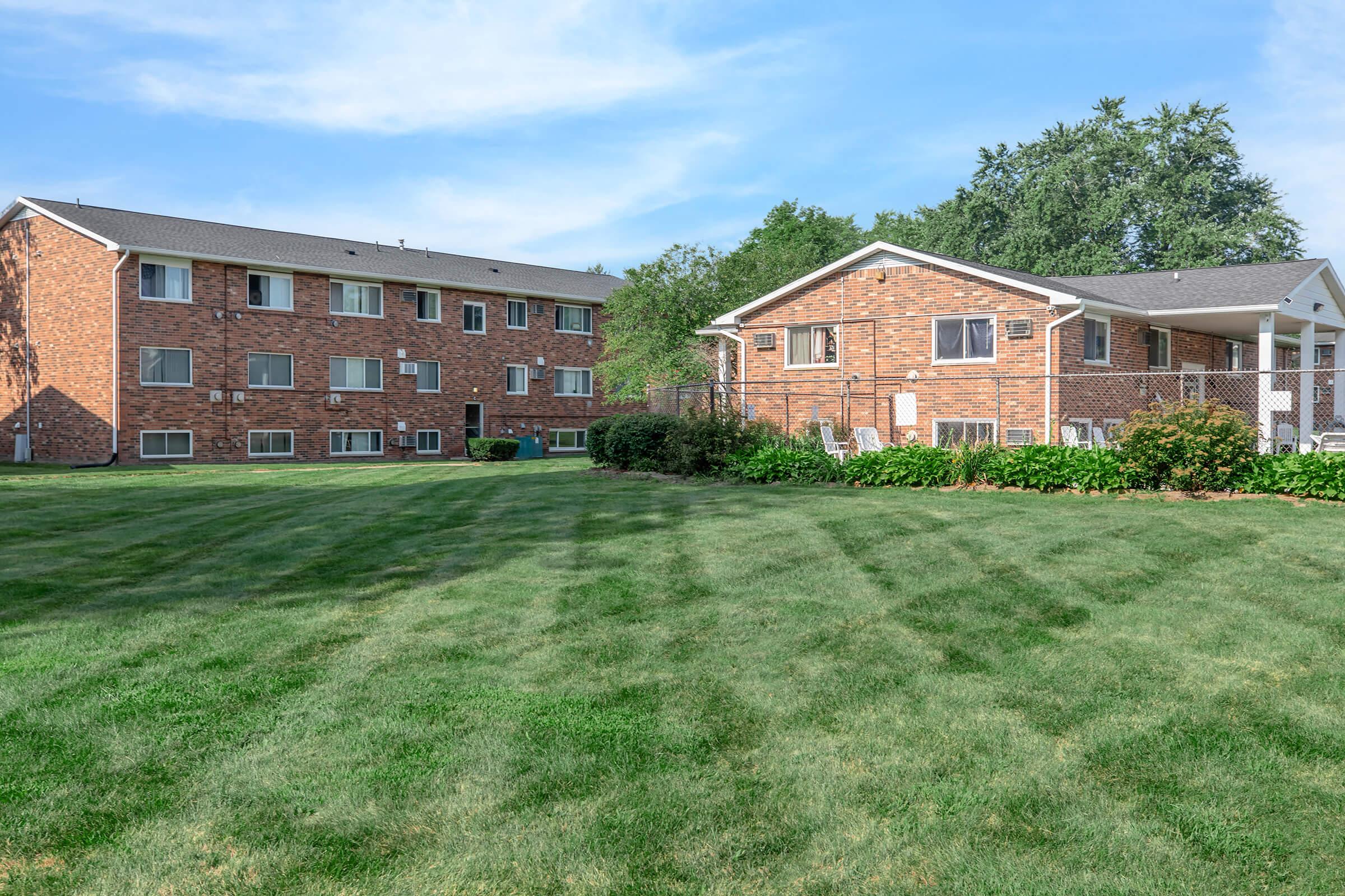 A grassy area with well-maintained lawns, featuring two brick buildings: a larger multi-story apartment complex on the left and a smaller single-story building on the right. The sky is clear with a few clouds, and there are trees in the background, creating a pleasant residential environment.
