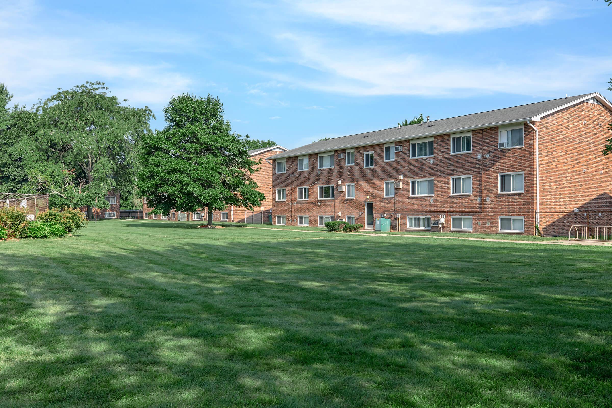 A view of a grassy area in front of a brick apartment building, featuring several windows. The scene is set under a clear blue sky with a few clouds and includes trees and manicured bushes in the foreground, creating a serene residential environment.