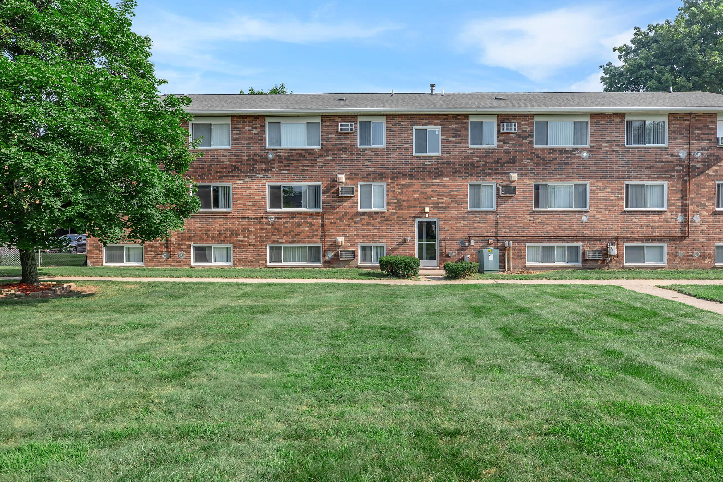 An apartment building with a brick facade, featuring multiple windows and air conditioning units. The front yard is grassy with a paved pathway leading to the entrance, surrounded by a few trees providing shade. Clear blue sky in the background.