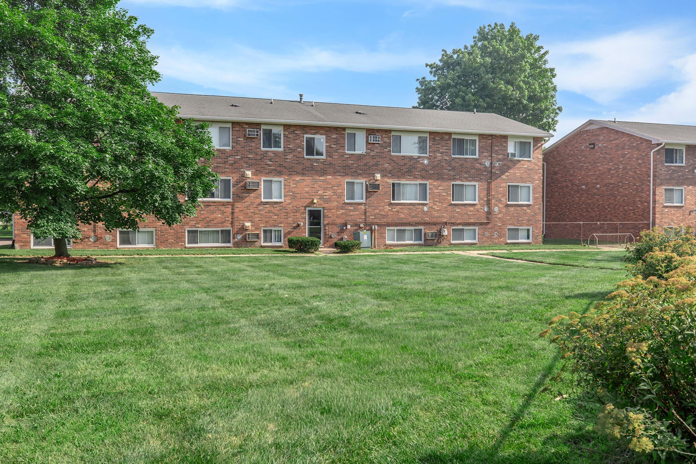 A view of a multi-unit brick apartment building surrounded by green grass and trees under a clear blue sky. The building features multiple windows and a well-maintained lawn in the foreground.