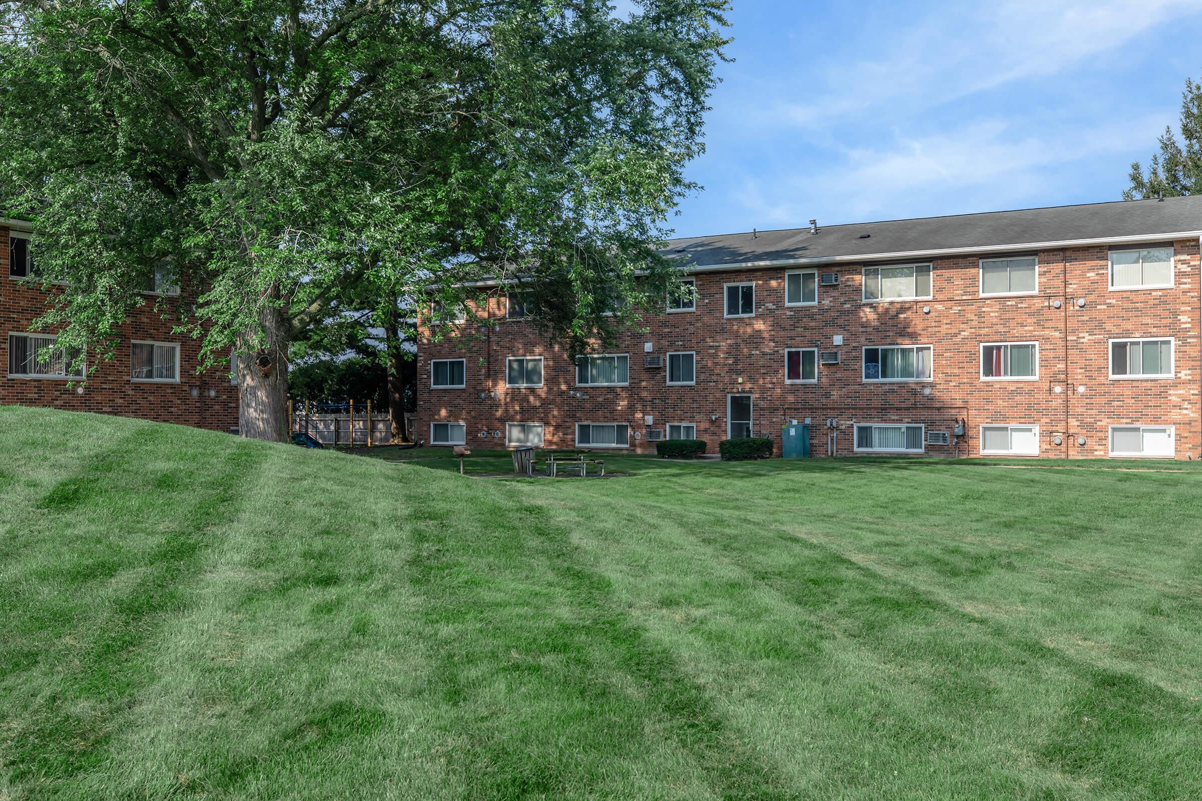 A grassy area with neatly mowed stripes in the foreground, bordered by two brick apartment buildings with multiple windows. A large tree provides shade, and there are a couple of benches visible on the lawn. The sky is clear with some clouds.