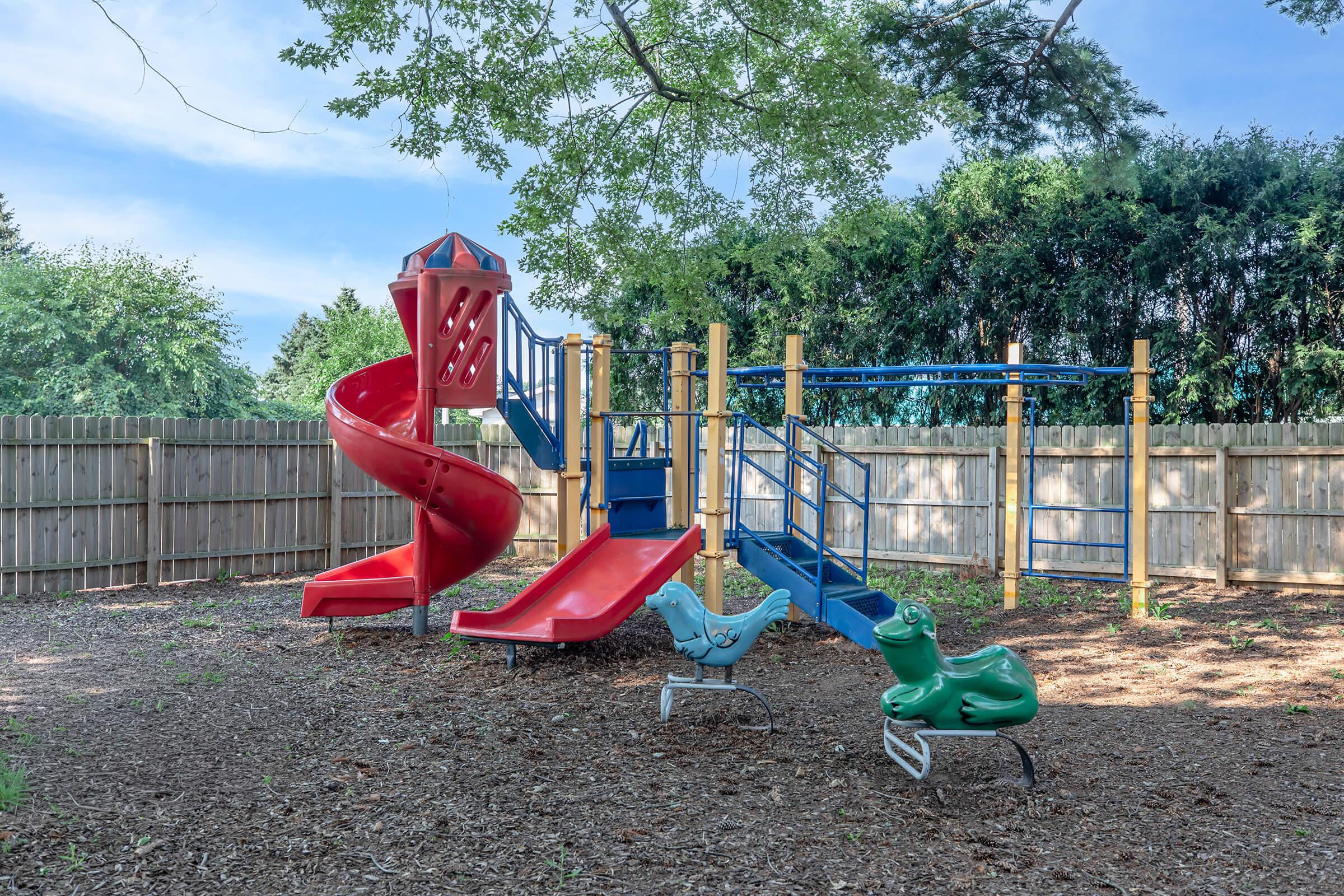 A children's playground featuring a red spiral slide, two red slides connected by a climbing structure, and two green rocking animals. The area is surrounded by a wooden fence and has a soft ground cover of mulch, with trees providing shade in the background.